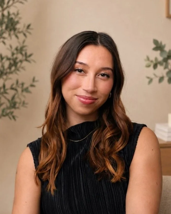 A young woman with long, wavy brown hair and a slight smile, wearing a sleeveless black top, sitting in a room with beige walls and green plants in the background.