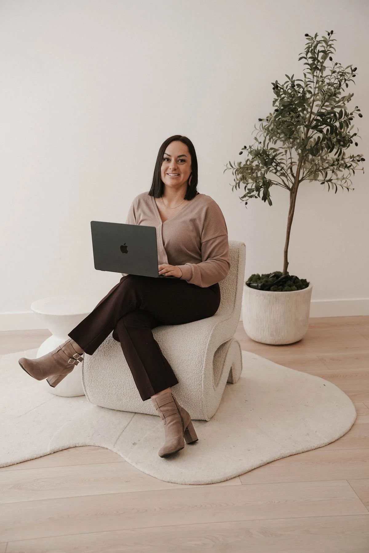 A woman with dark hair sitting on a cream-colored modern chair, holding an Apple MacBook, in a minimalistic room with light wood flooring, a small white side table, a white rug, and a tall potted plant in the background.