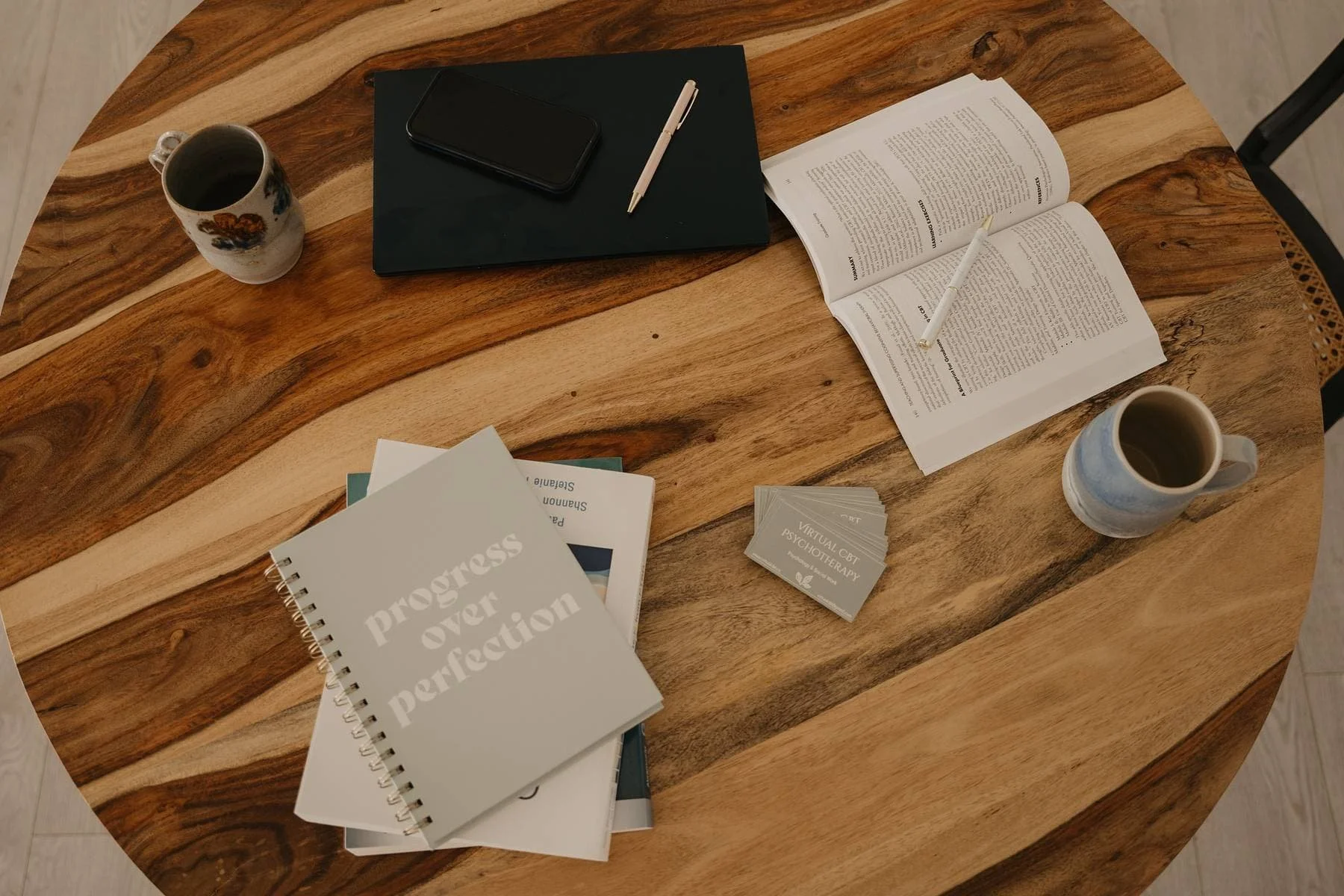 A wooden table with notebooks, magazines, a smartphone, a pen, two coffee mugs, an open book with a candle, and a stack of business cards.