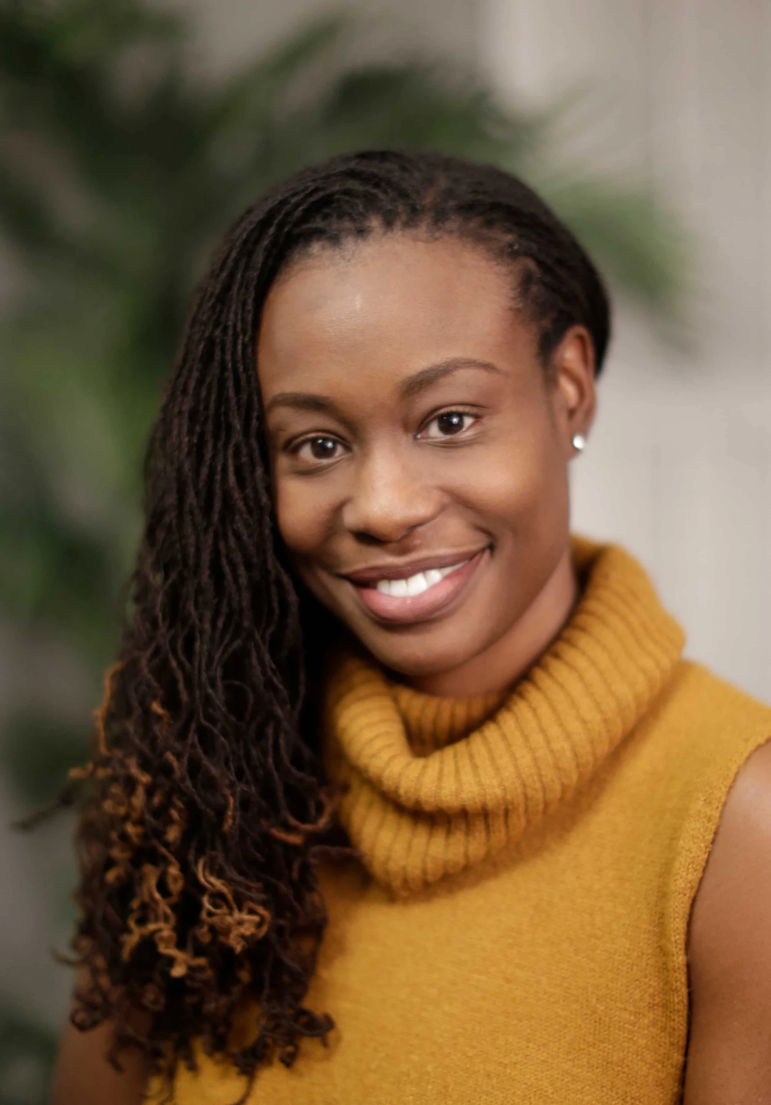 A woman with dark skin, long styled hair, and a warm smile, wearing a mustard yellow turtleneck sweater.