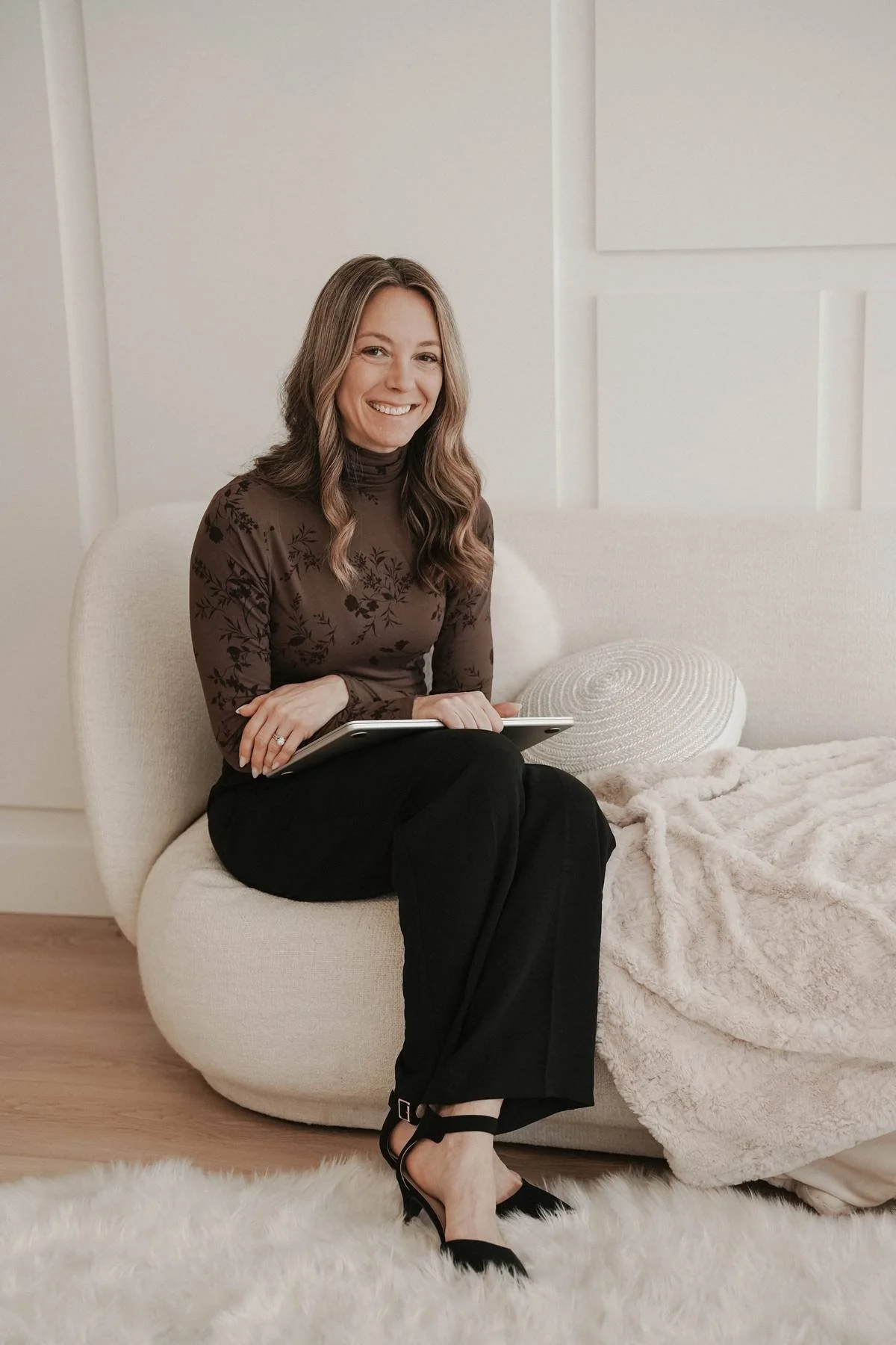 Woman with long wavy hair smiling at the camera, wearing a brown turtle neck and black dress pants. Holding a laptop and sitting on a white couch