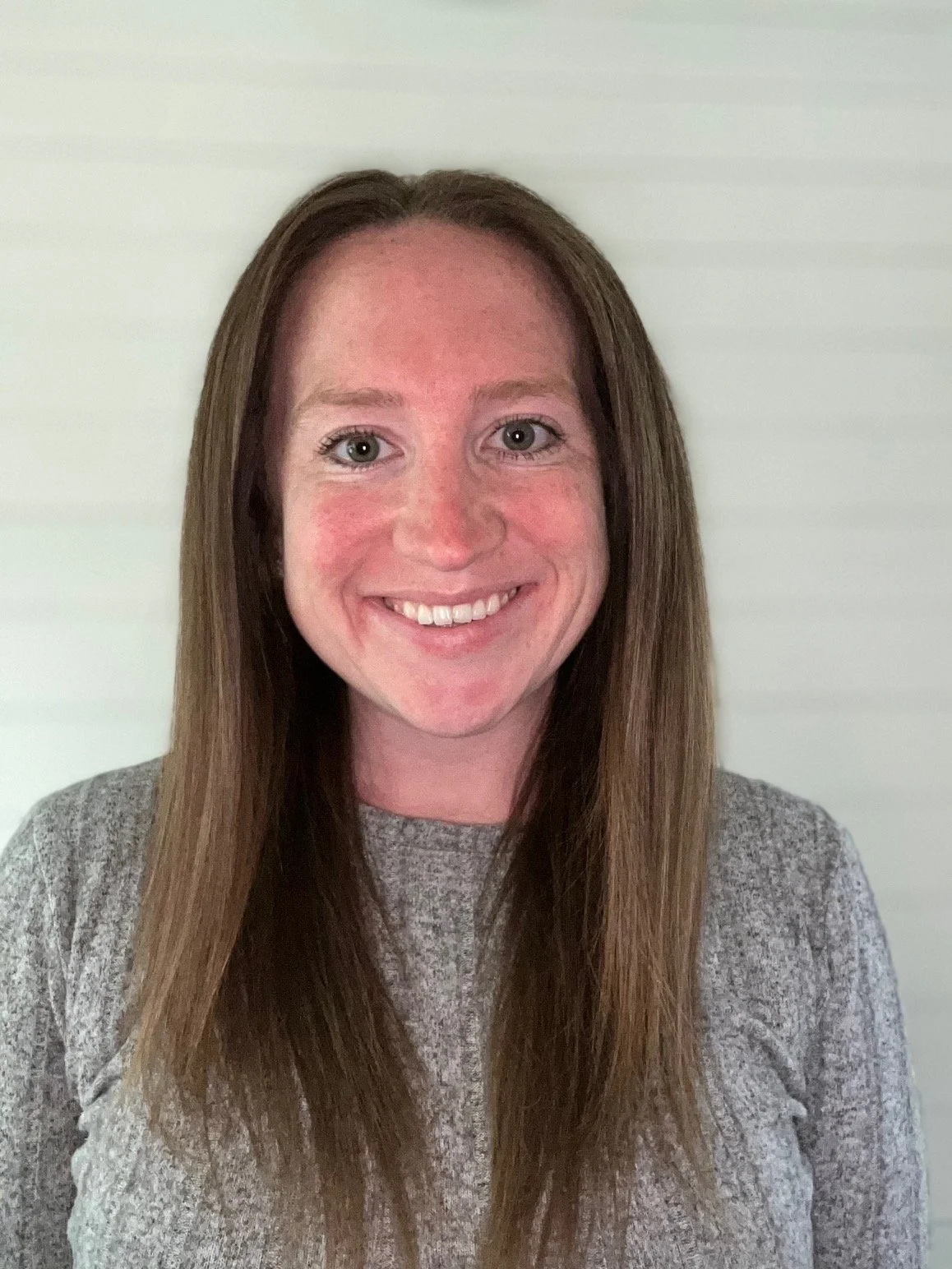 A woman with long brown hair, smiling, wearing a gray sweater, standing against a light-colored wall.