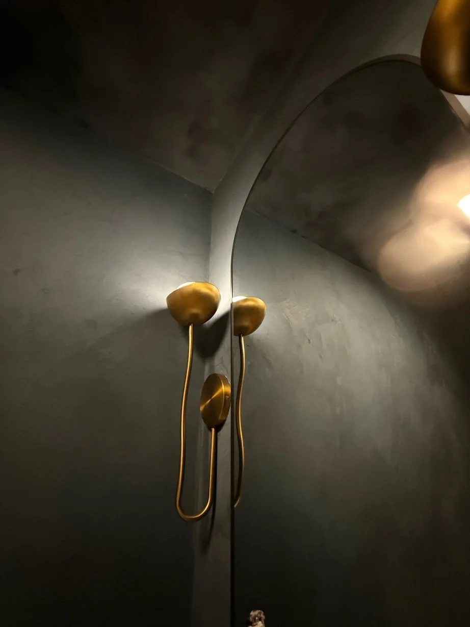 Close-up of a wall-mounted modern lamp with a curved metallic arm and three irregularly shaped, golden light fixtures against a dark wall.