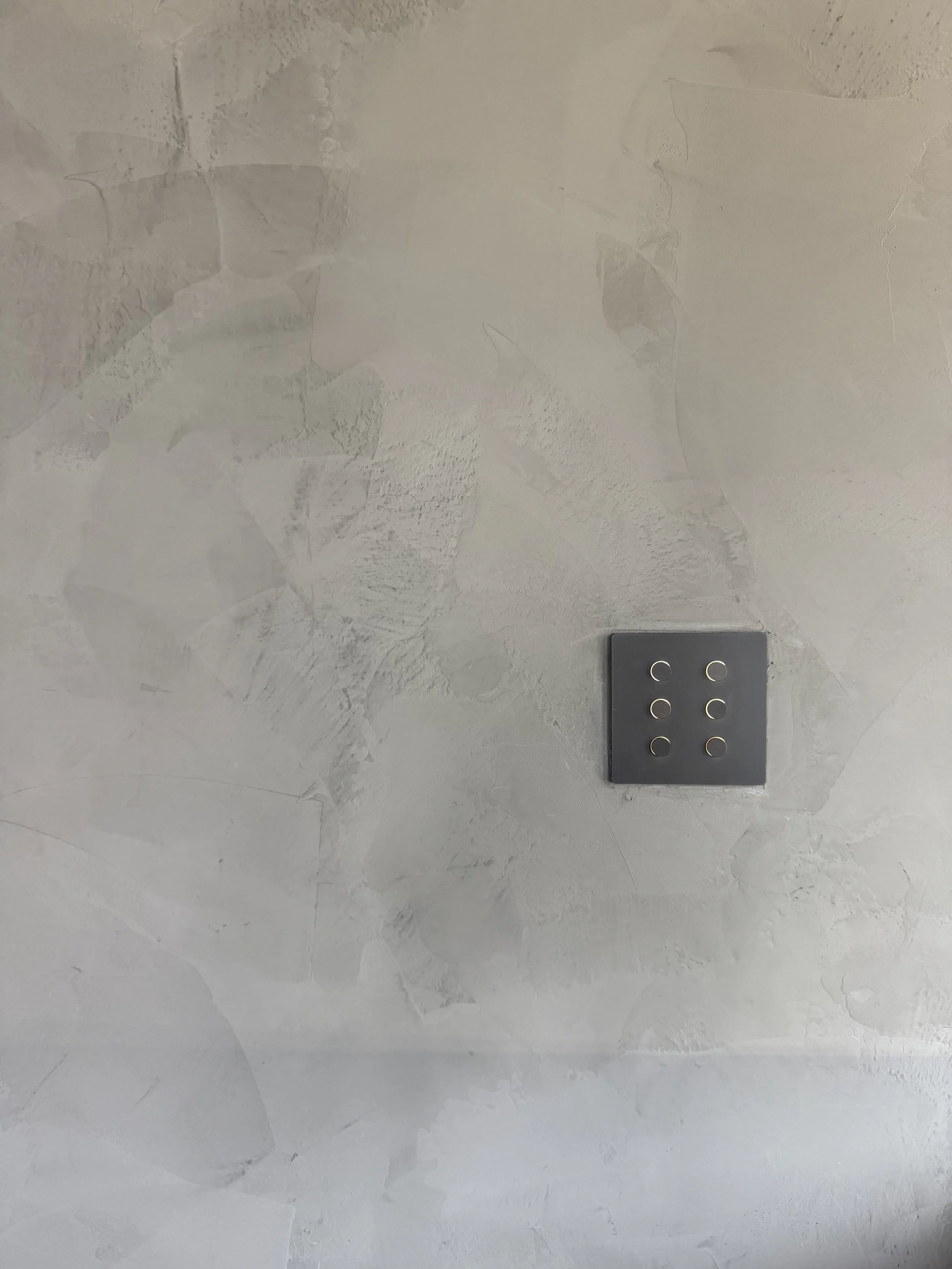Light gray textured wall with a gray square electrical switch plate containing six small circular switches.