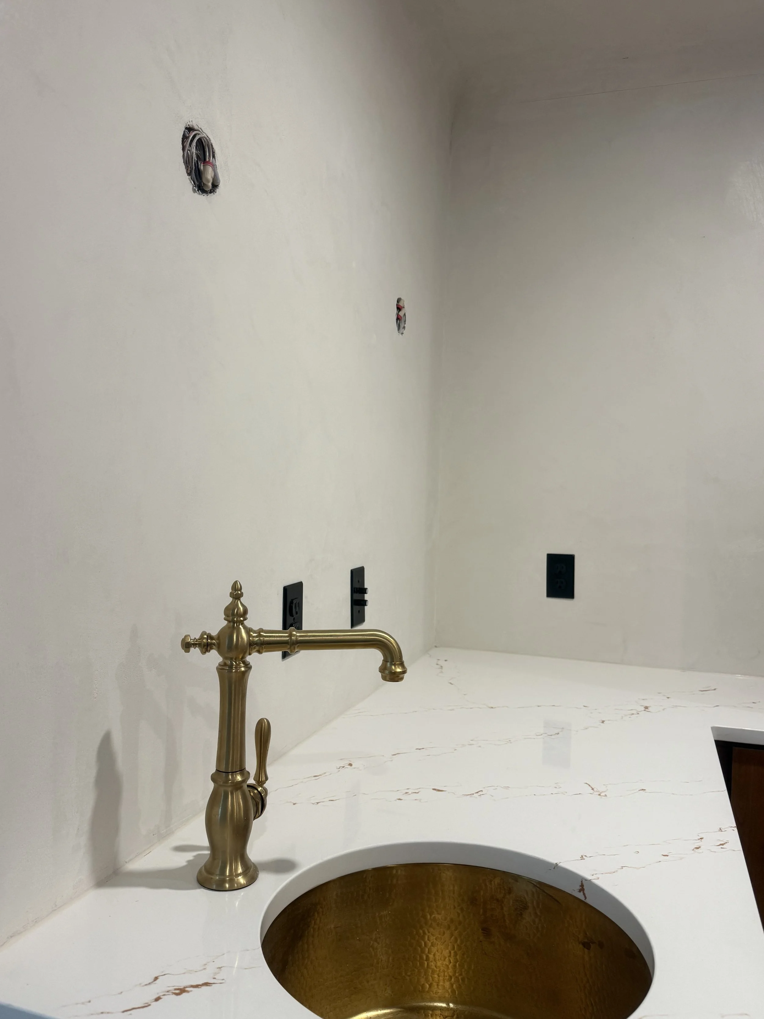 A kitchen countertop with a brass sink and matching faucet, some wall electrical outlets, and unfinished electrical wiring in the wall.