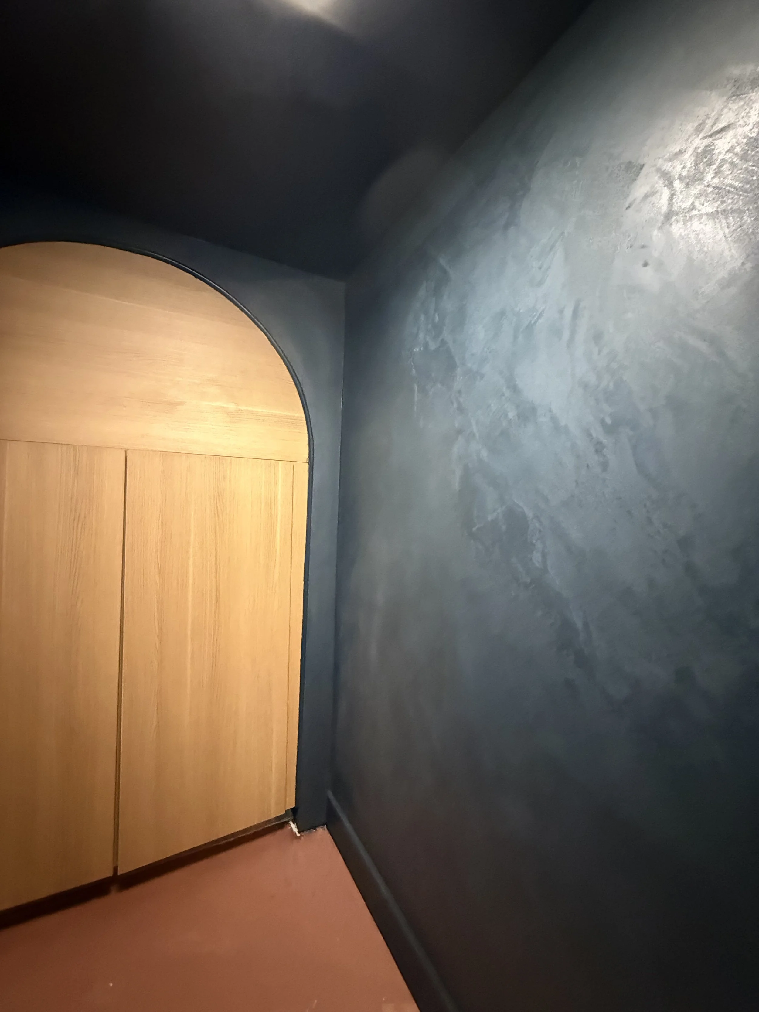 Interior corner with a dark blue wall, wooden cabinet with rounded top, and a reddish-brown floor.