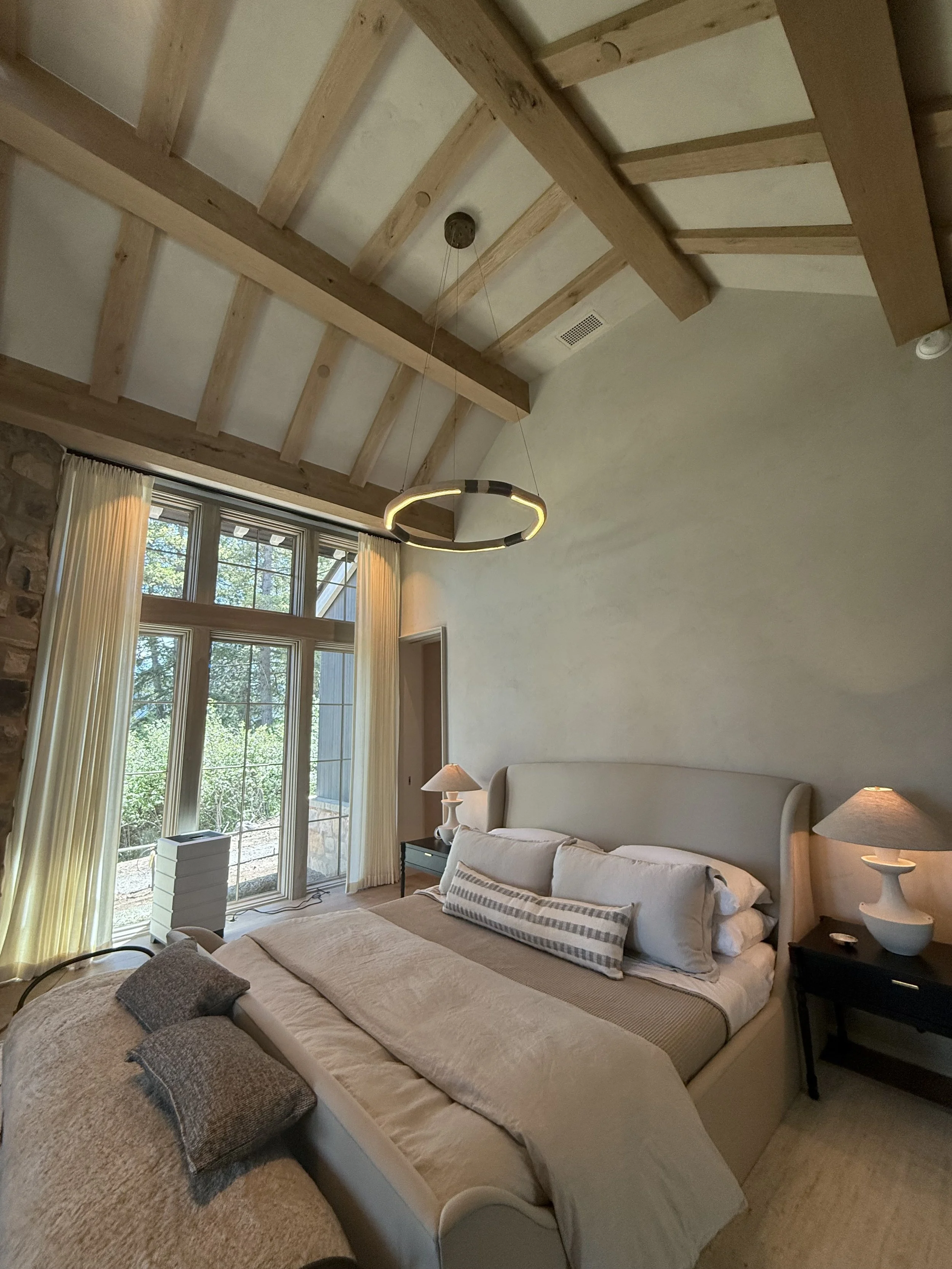 A bedroom with a high vaulted ceiling featuring wooden beams, a modern circular chandelier, a large window with cream curtains, a beige upholstered bed with white and striped pillows, and two black nightstands with lamps.