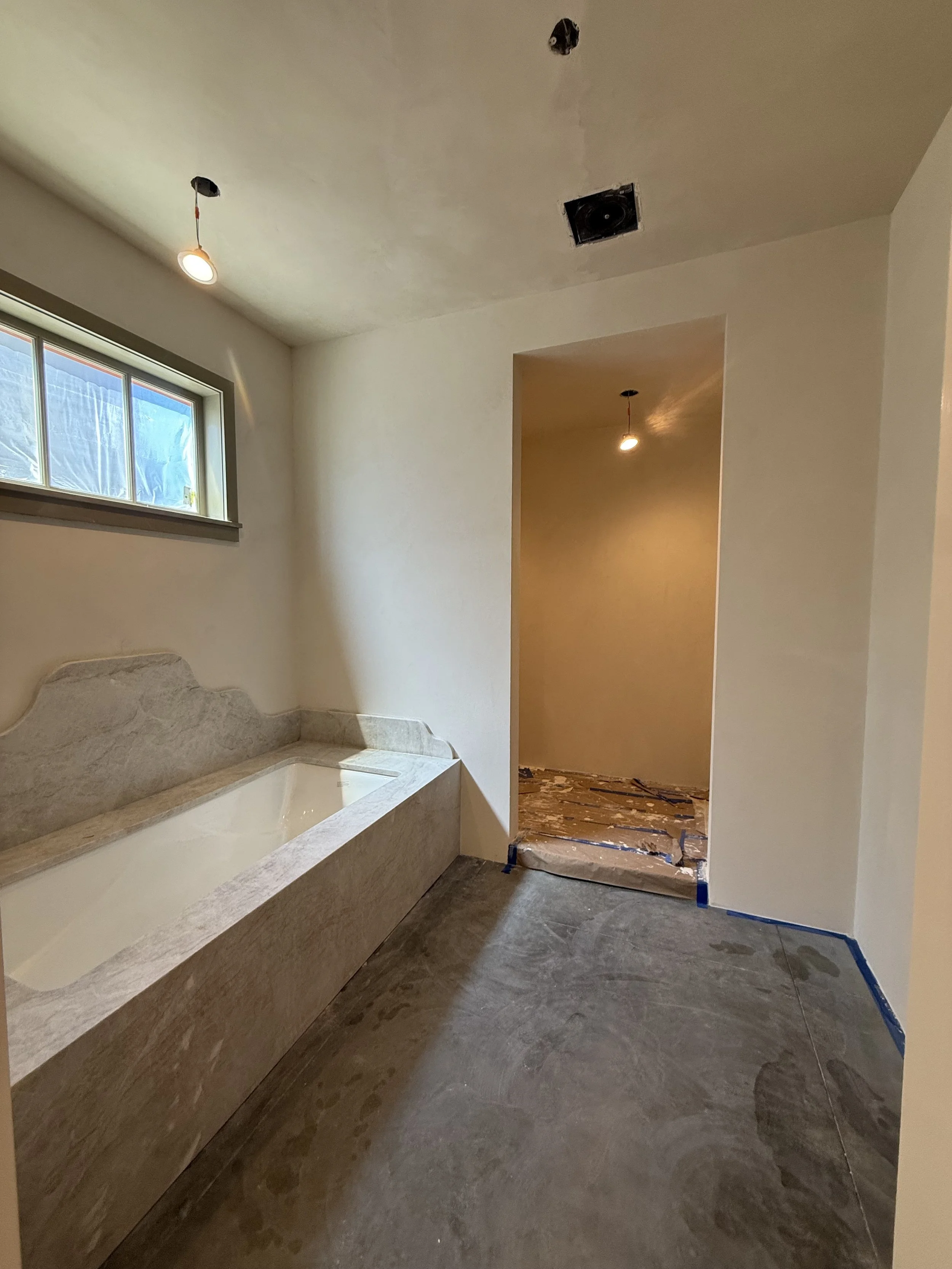 Bathroom under renovation with a bathtub made of stone, a small window, and a partially finished ceiling with exposed light fixtures and ductwork.