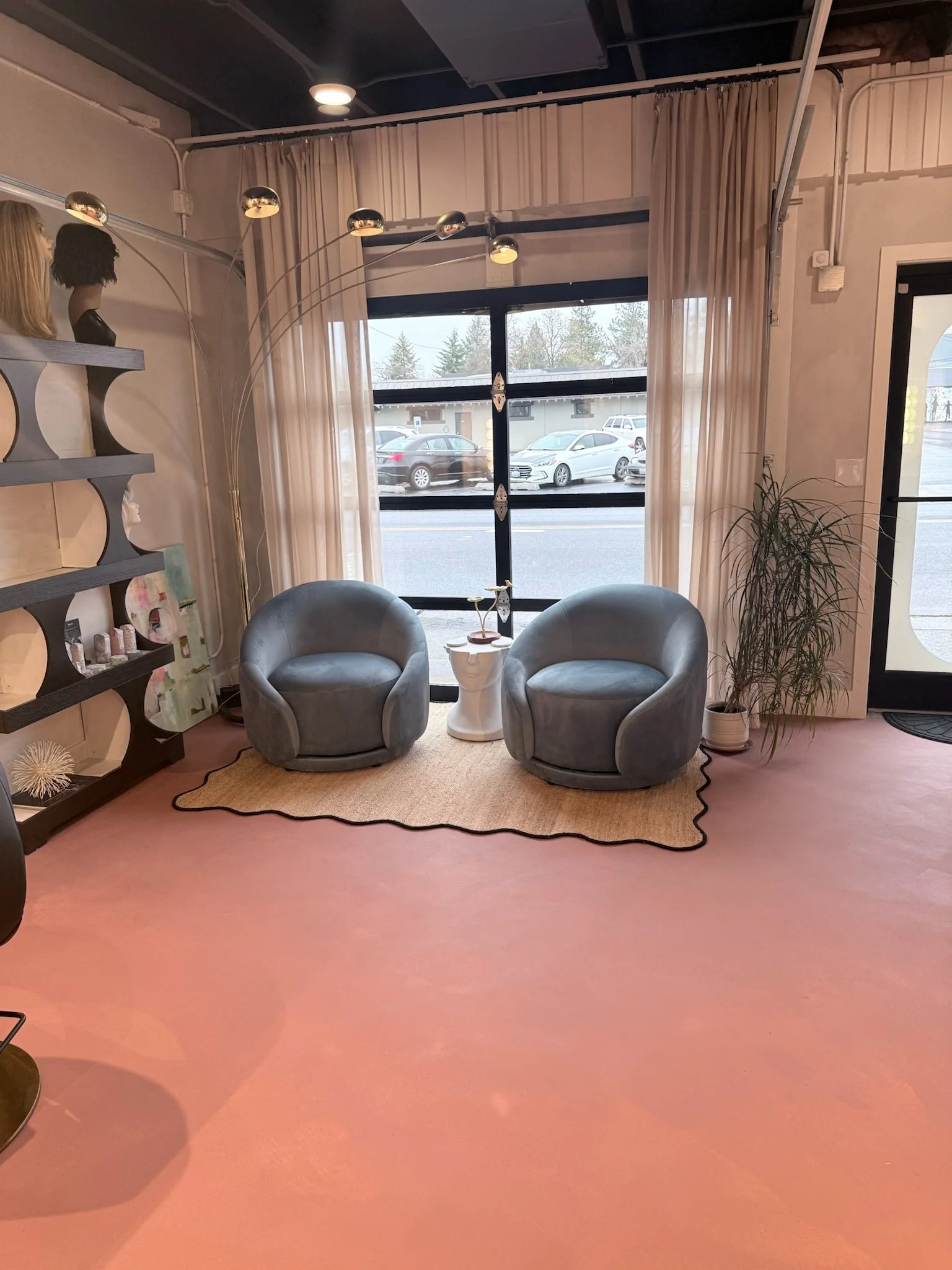 Interior of a modern salon or waiting area with two gray armchairs, a small white side table between them, a beige rug, a tall potted plant, a large window with white curtains, a black door to the right, and a multicolored overhead light fixture.