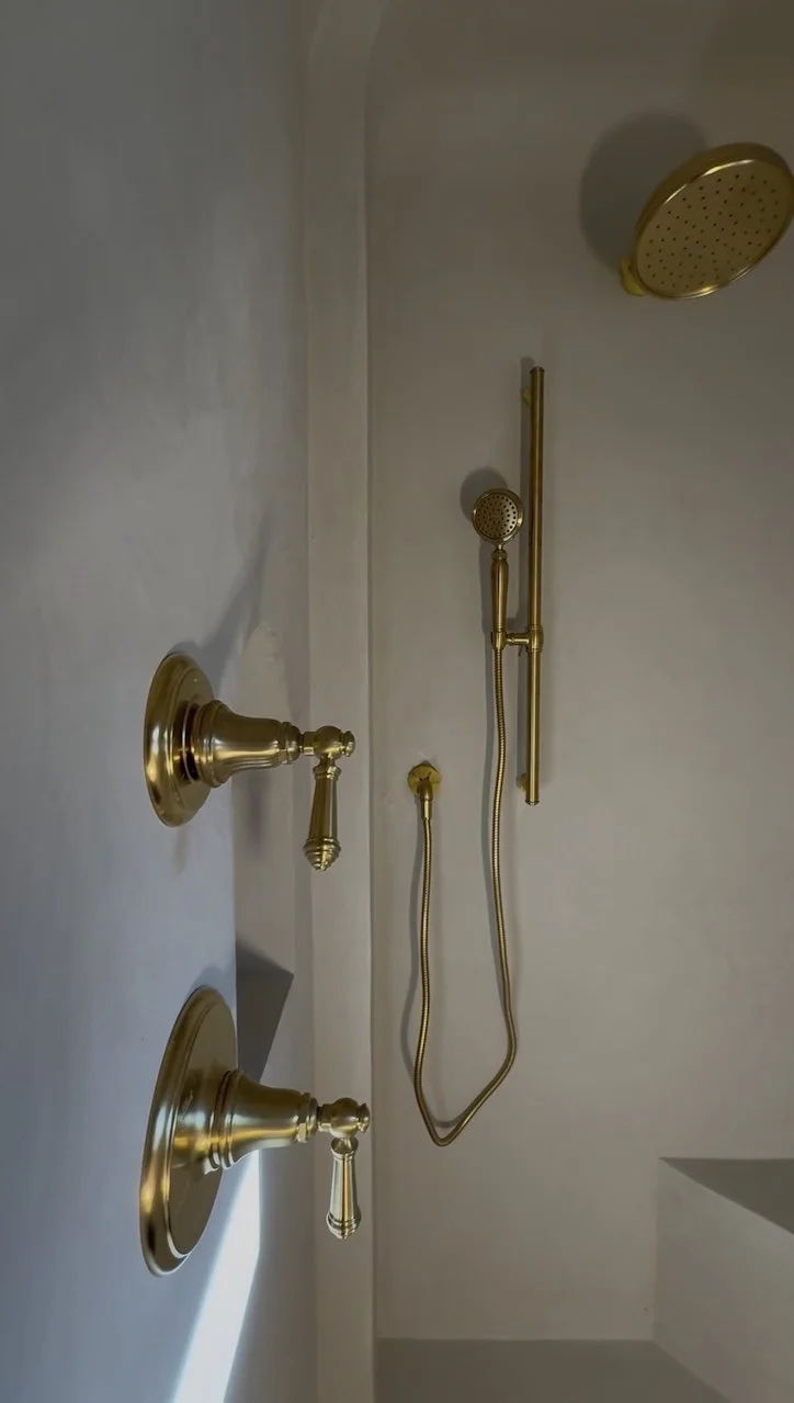 Wall-mounted shower with gold fixtures, including a large overhead showerhead, a handheld showerhead, and two thermostatic mixer controls.