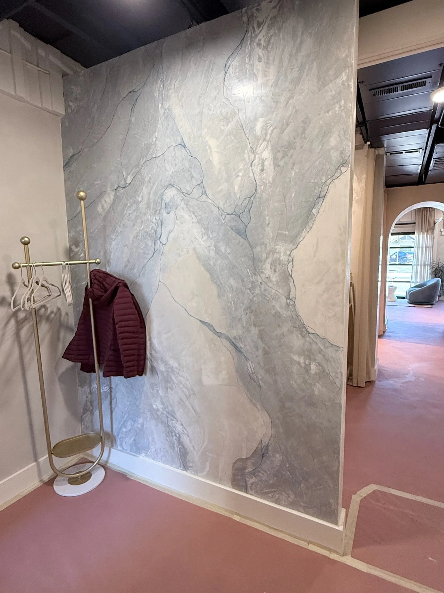 A corner of a room with a large marble-patterned wall, a pinkish carpeted floor, a gold coat rack with a burgundy jacket, and an arched doorway leading to a lounge area with a light grey chair and window.