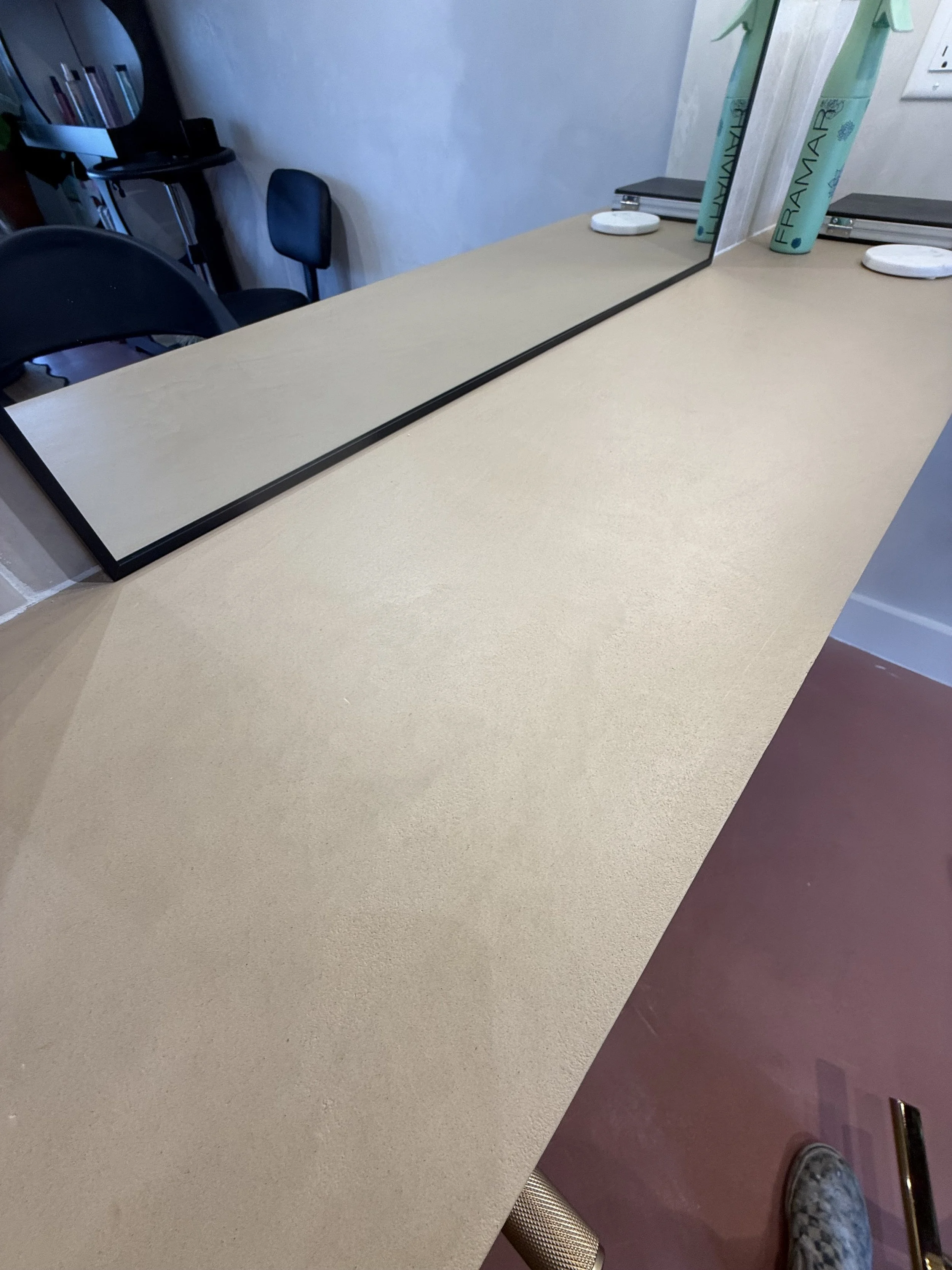 A beige countertop with a large mirror on it, reflecting part of the room and a green bag with "FRAMAR" printed on it. In the background, there are black chairs and a blue wall with some shelves.