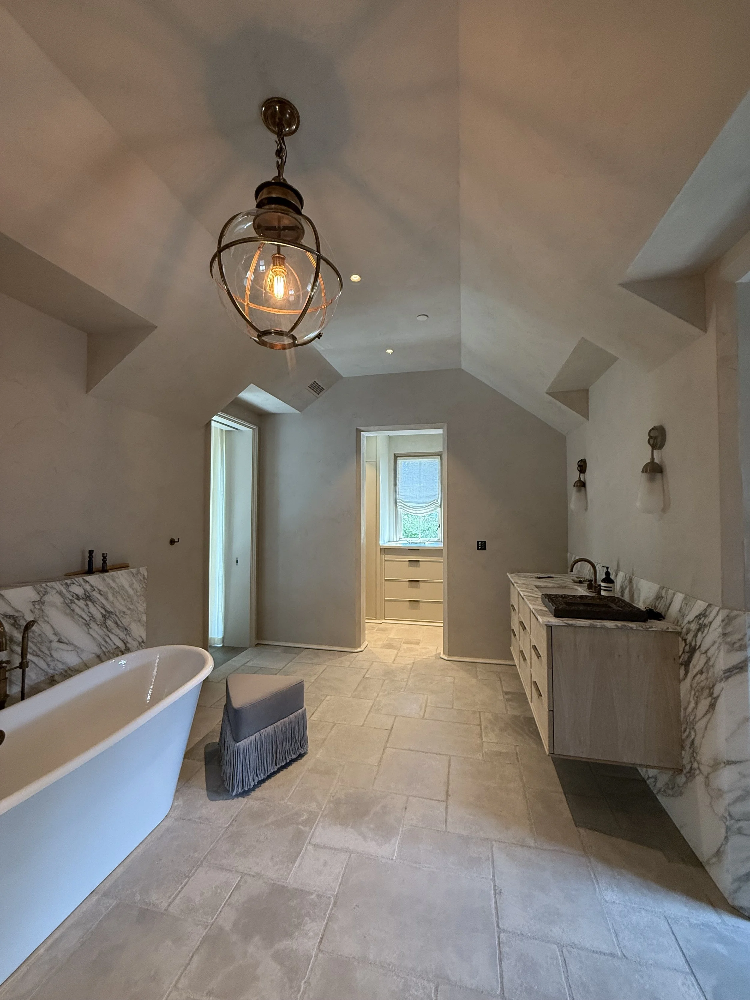 A spacious, modern bathroom with a freestanding bathtub, beige tiled floor, marble accents, and vanity with double sinks, illuminated by vintage-style pendant lighting and wall sconces.