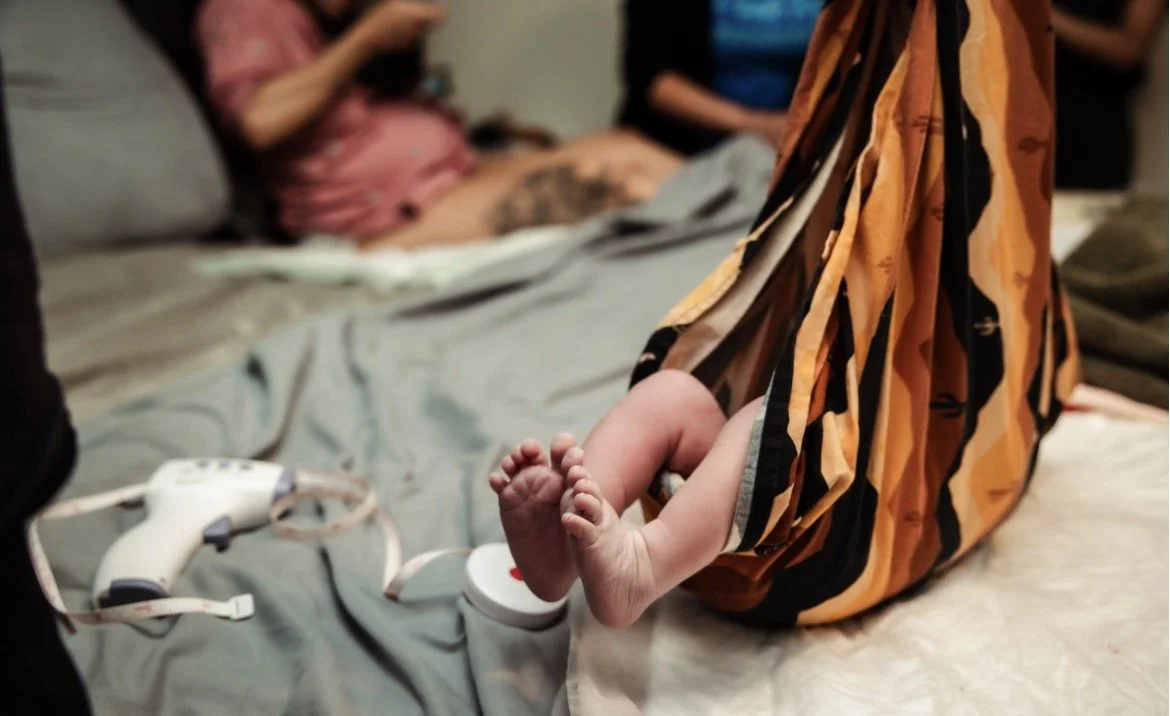 A newborn baby's feet sticking out from under a colorful patterned blanket on a bed. In the background, a person is sitting up in bed, wearing a pink shirt and looking at their phone. Medical equipment and monitors are visible. Homebirth