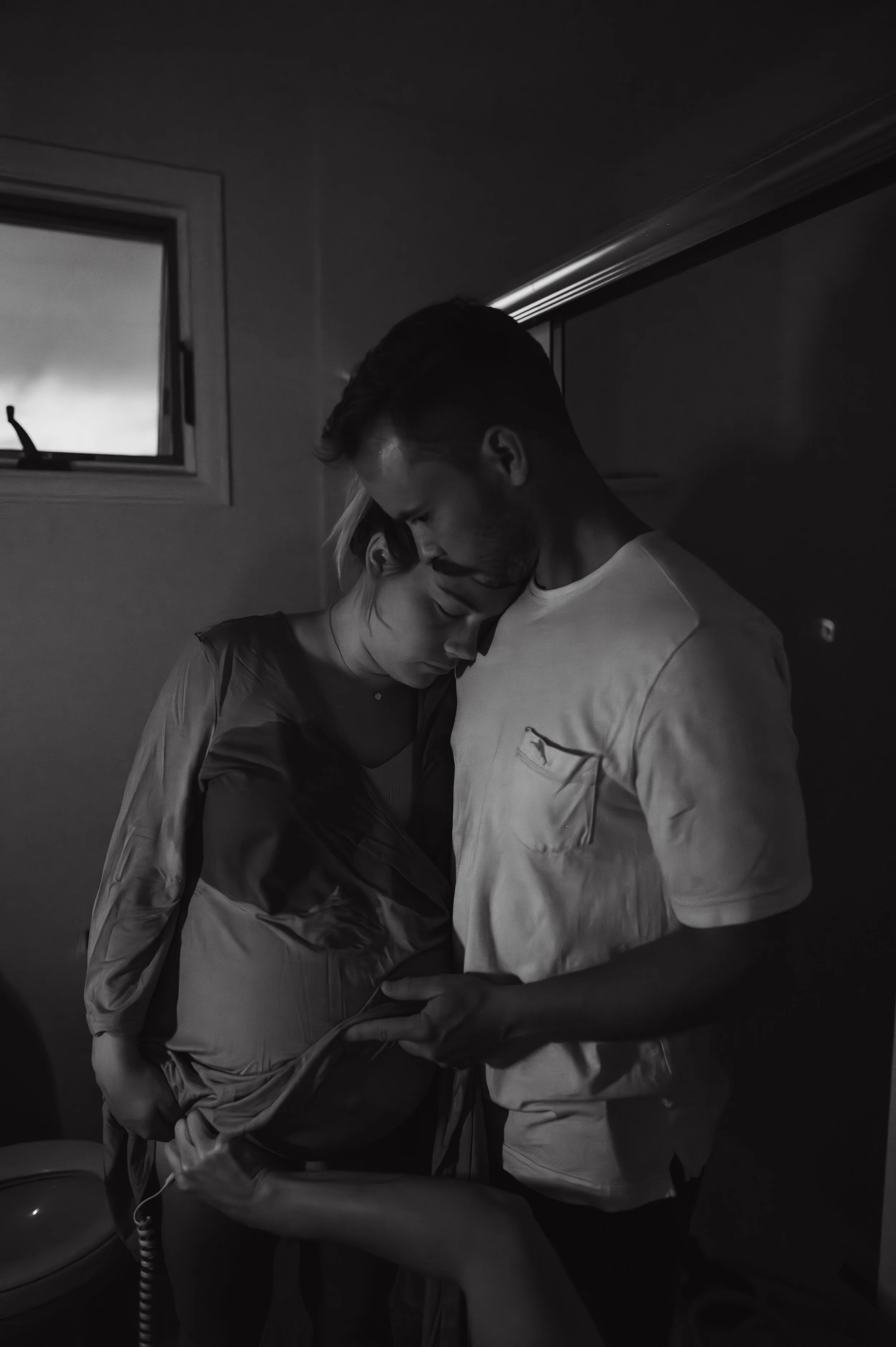 A black and white photo of a man and woman embracing, with their foreheads touching, in a dimly lit room near a window.