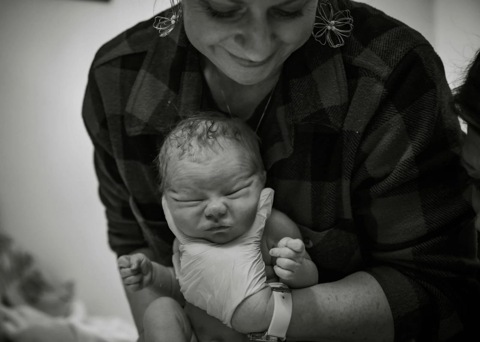 A newborn baby with curly hair and closed eyes is being held by a woman with a checkered shirt. The woman is smiling slightly and wearing butterfly earrings. The baby is wearing a diaper and is being gently supported by someone wearing a glove.