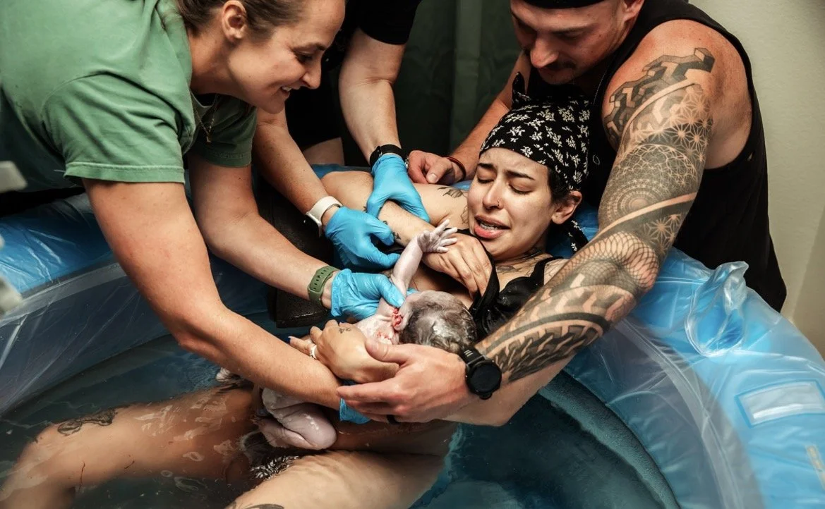 A woman giving birth in a hospital setting with medical staff assisting, holding a newborn baby.