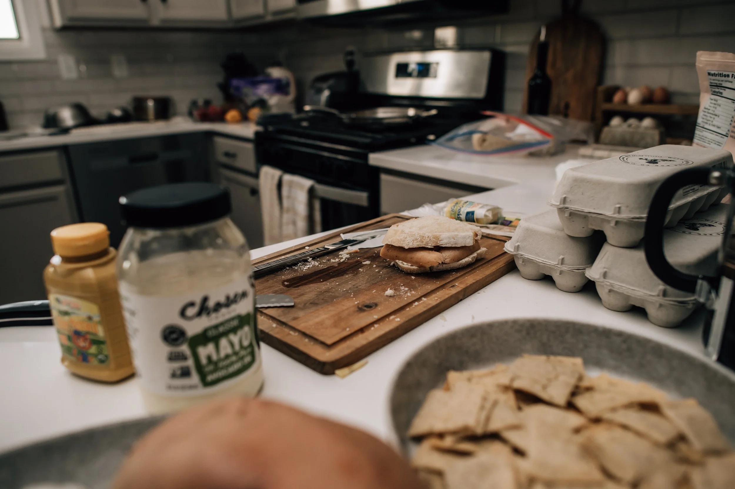 A cluttered kitchen counter with eggs in cartons, mustard and mayonnaise jars, a cutting board with sandwich ingredients, and a stove in the background.