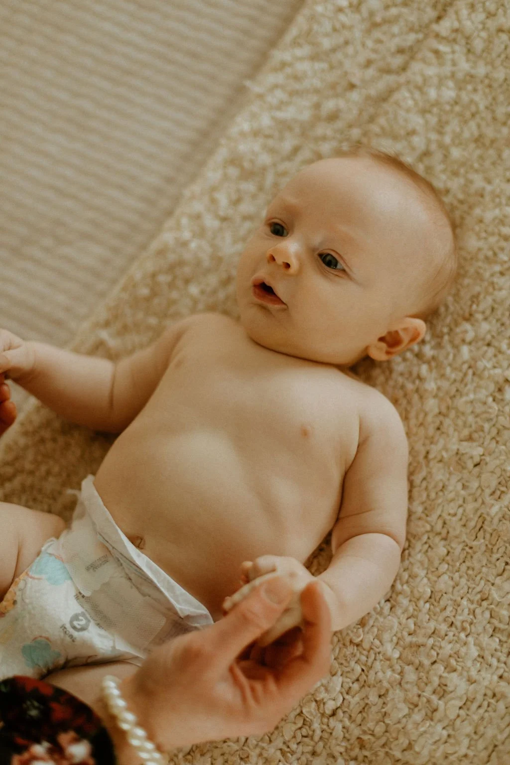 A baby lying on a beige textured blanket, wearing a diaper, holding an adult's hand, with a curious expression on the face.