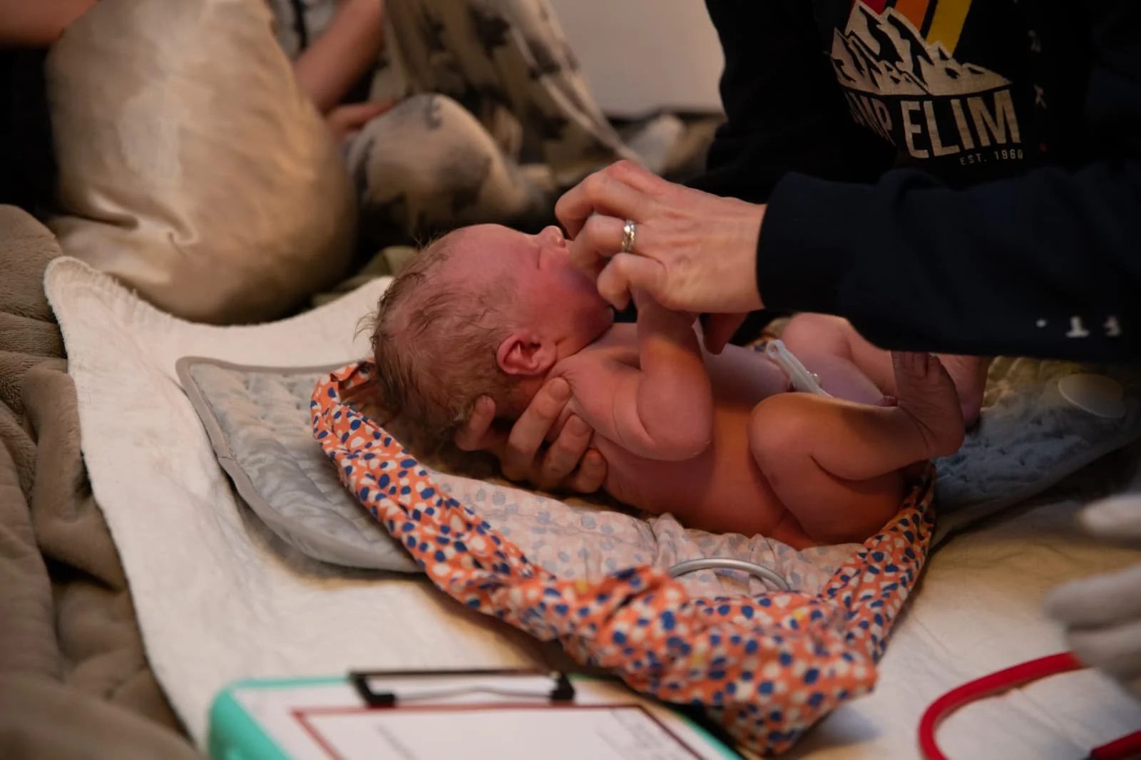A newborn baby lying on a blanket, being cradled and touched by a person in a dark shirt, in a hospital or medical setting.