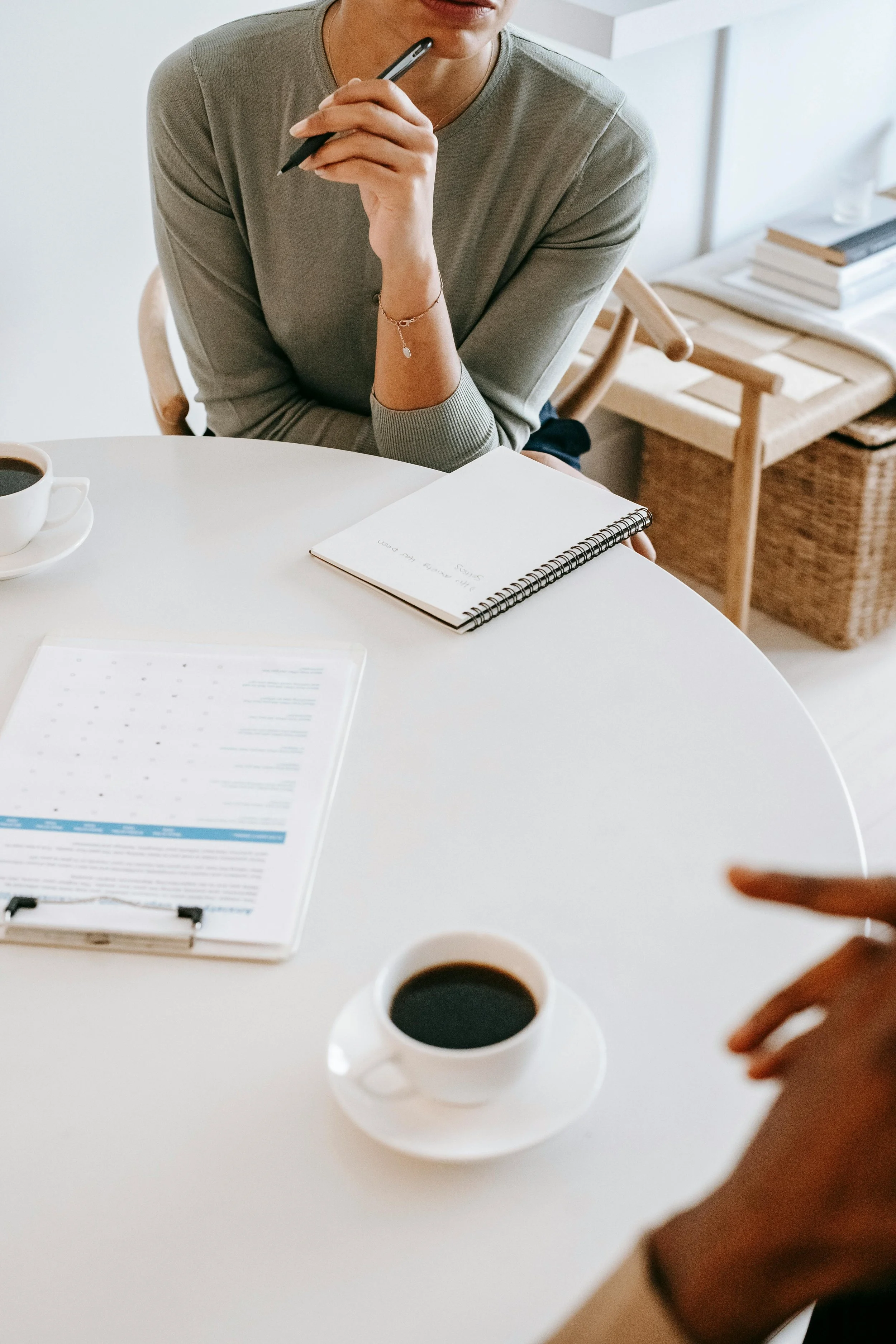 A close up image of a white desk with a woman seated with a notepad and cup of coffee