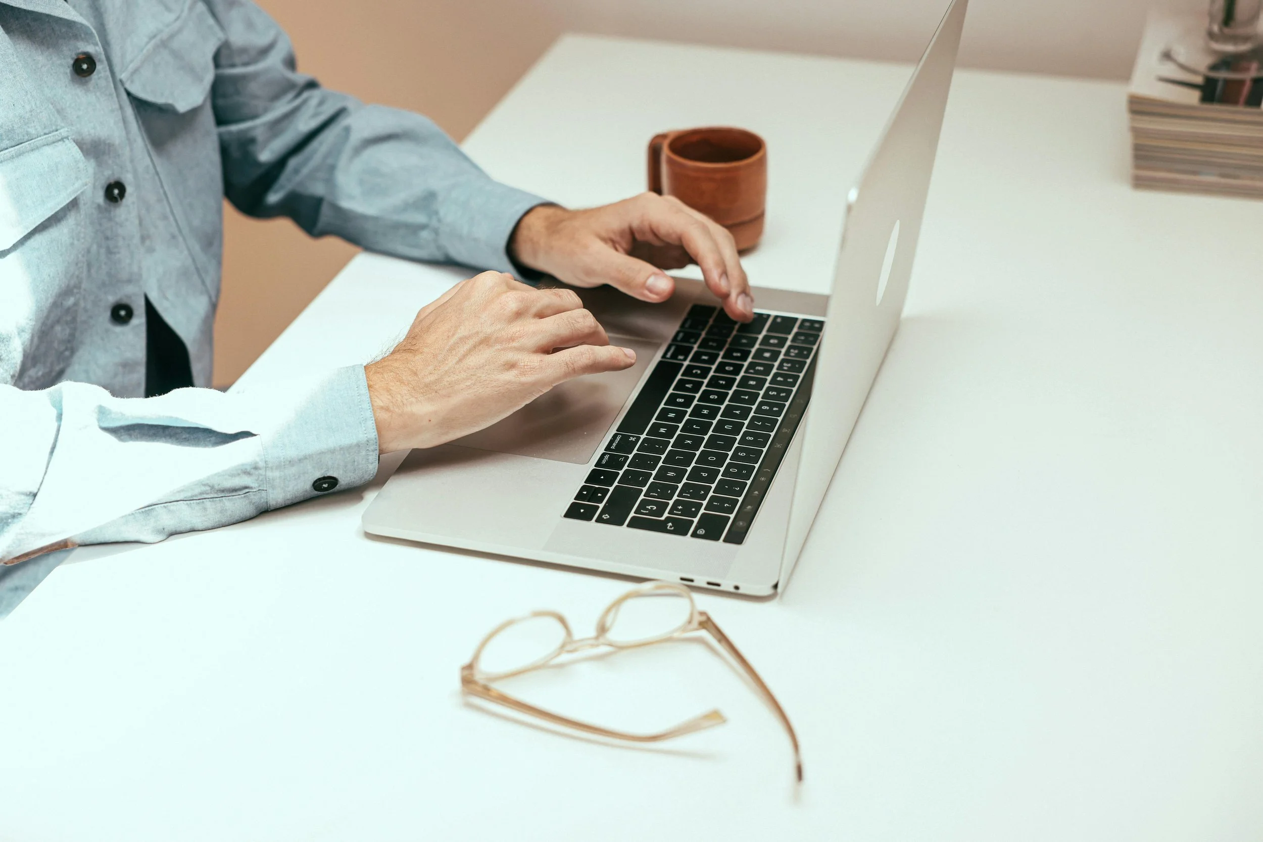 A man's hands typing on a laptop next to a mug and a pair of glasses sitting on a white desk
