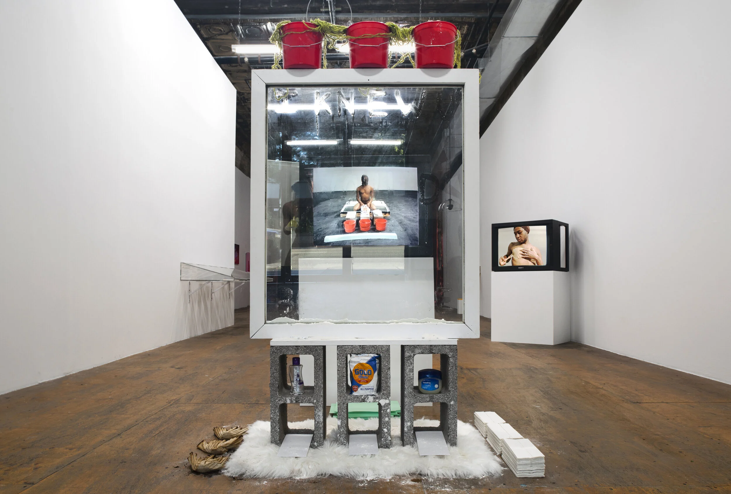Window View: Covered in Lube, 2014–16, 2021, in&nbsp;Keioui Keijaun Thomas: Hands Up, Ass Out, curated by Shehab Awad as Executive Care*, 2021 at Participant Inc, New York. Photo: Daniel Kukla.Visual Description: A towering sculpture made up of a large, single-pane window stands atop three concrete cinder blocks. Three red, plastic buckets with gold yarn sit on the window’s top ledge. The words “I KNEW U WOULD BE HERE” are inscribed in lube on the glass panel. A monitor playing a video is visible through the glass.
