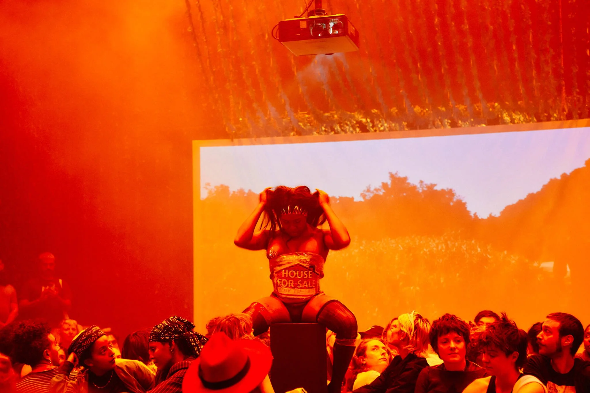 Artist is lit with bright red, seated on a black block adjusting her unit surrounded by audience.