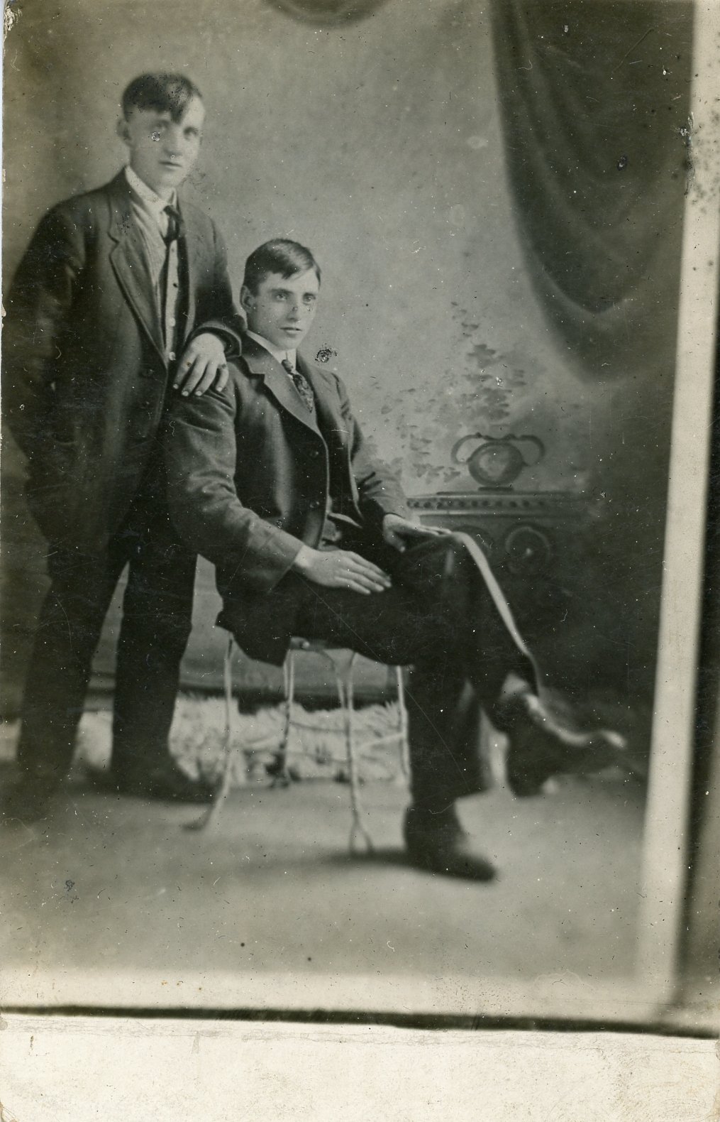 Vintage black and white photograph of two men in suits posed in a formal studio setting, one standing and the other seated.