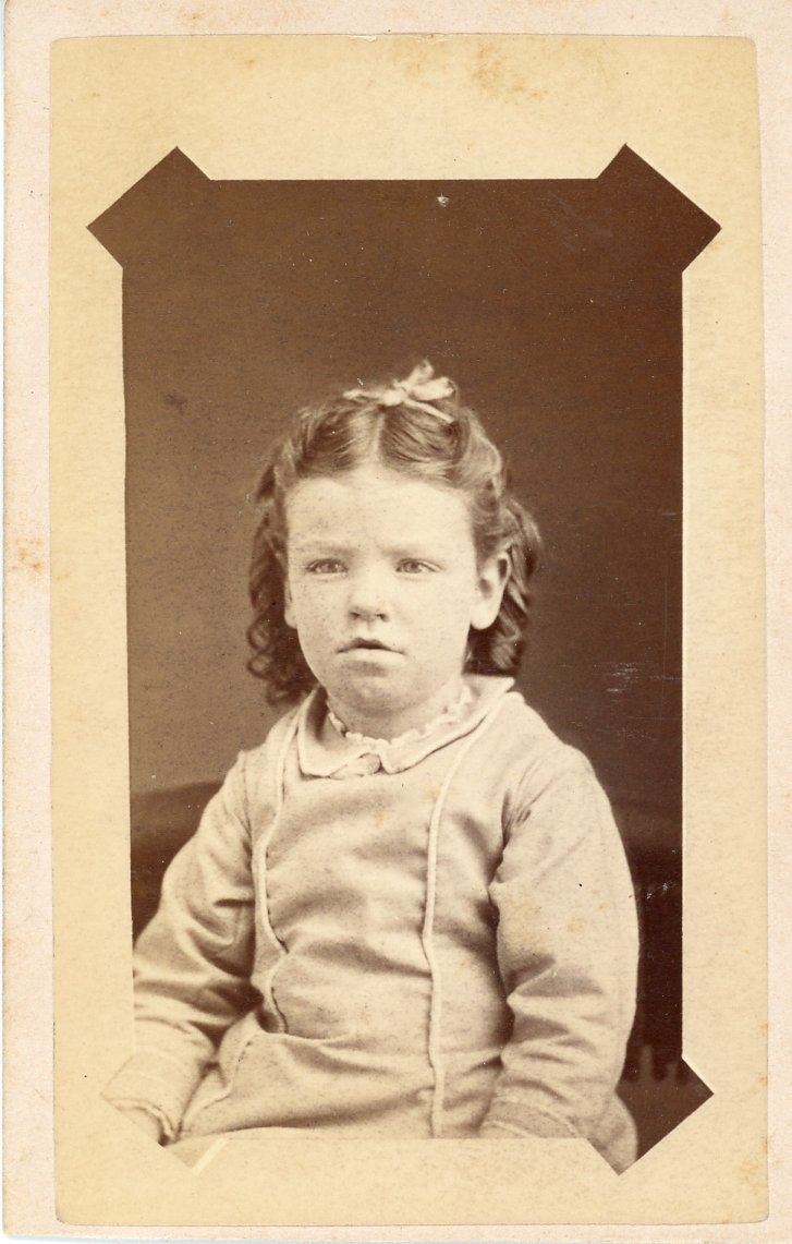 Vintage portrait of a young child with curly hair wearing historical clothing. The photo is framed in a cabinet card style.