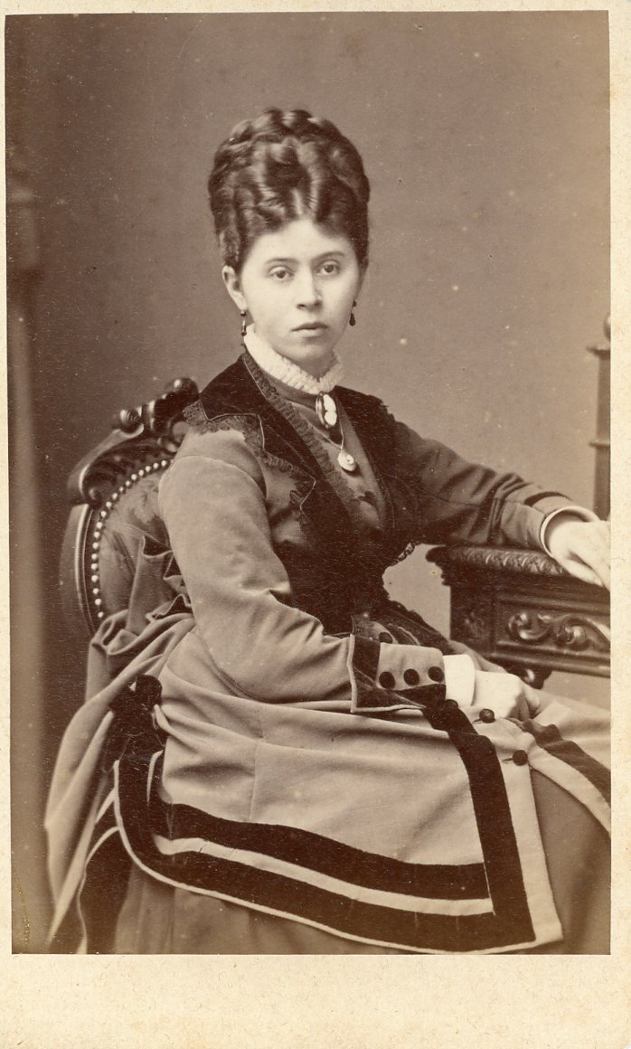 Victorian-era woman seated, wearing a formal dress with intricate patterns, in a vintage studio setting.