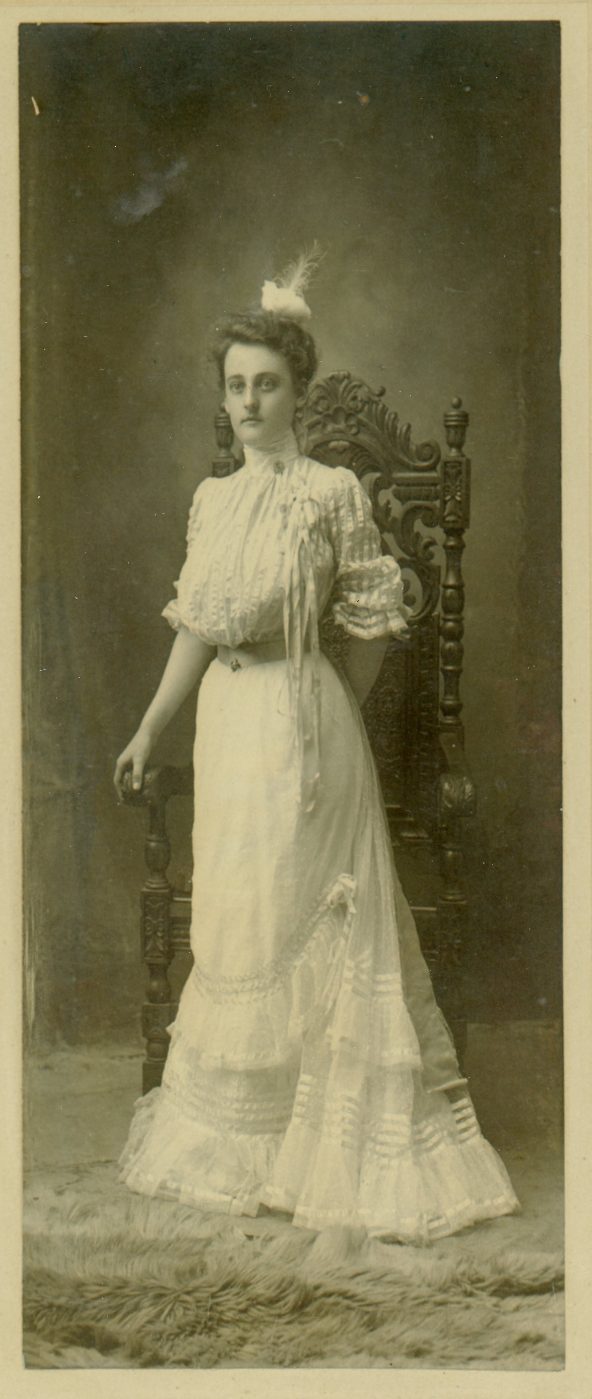 Vintage photograph of a woman in an ornate chair, wearing a white Victorian dress, circa early 20th century. She stands with a serious expression, showcasing intricate lace details on her dress with a feathered hair accessory.