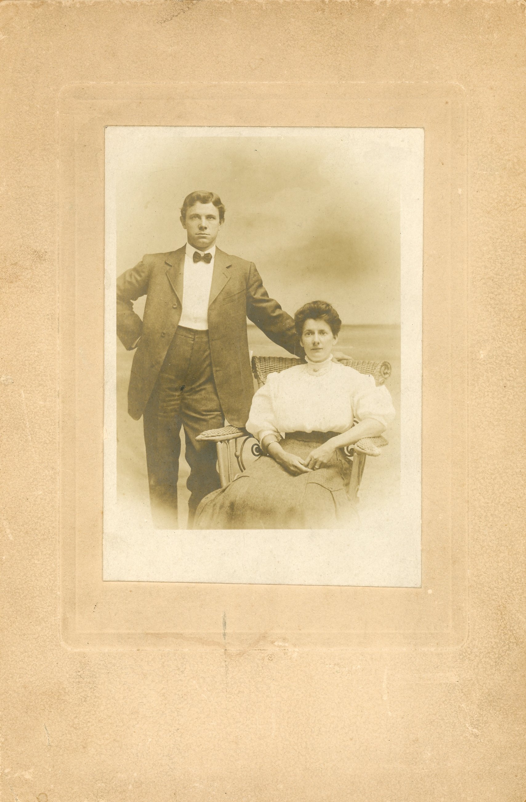 Vintage sepia photograph of a man in a suit standing beside a seated woman in a formal dress, on a textured background.