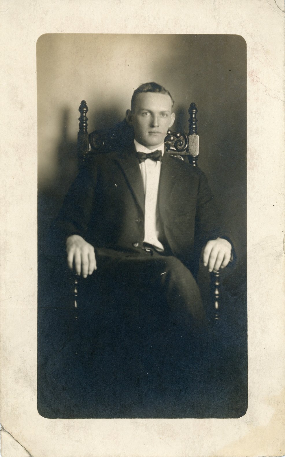 Vintage portrait of a man in a suit with a bow tie, sitting on a decorative chair.
