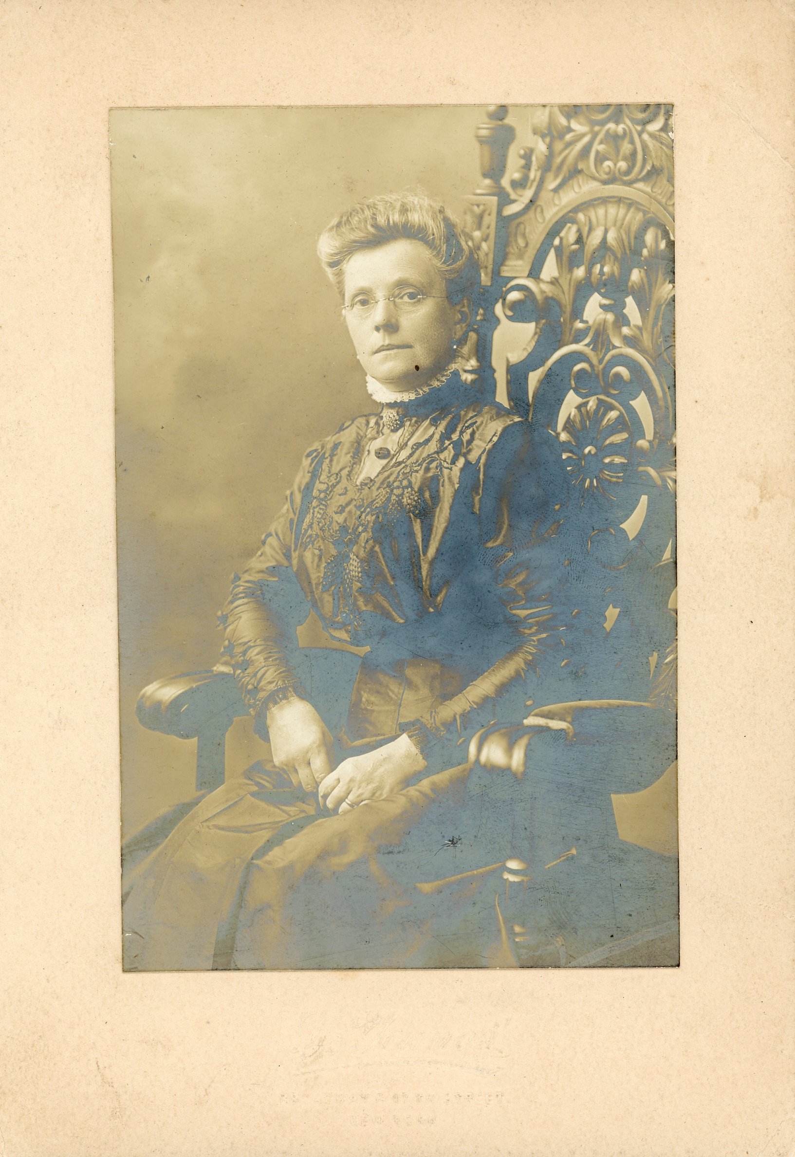 Vintage photograph of a woman seated in an ornate chair, wearing early 20th-century attire with lace details and glasses, framed on a textured paper background.
