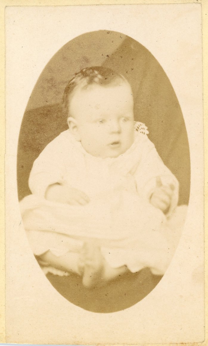 Sepia-toned vintage photograph of a baby wearing a white dress and sitting, with a soft-focus background.