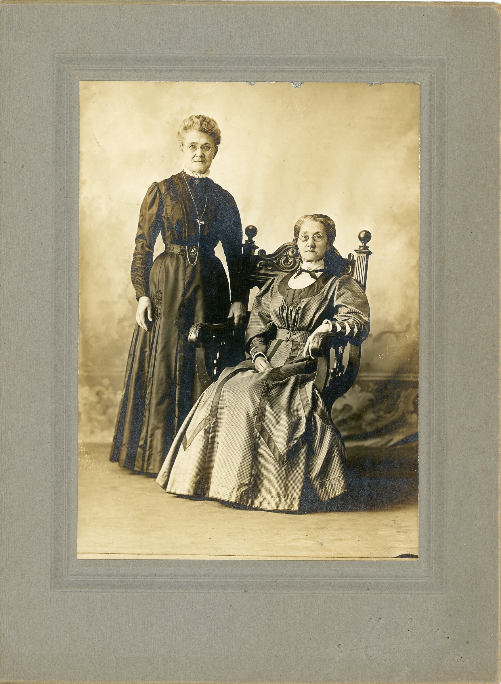 Vintage photograph of two women in formal attire, one standing and the other seated in an ornate chair, with a neutral backdrop.