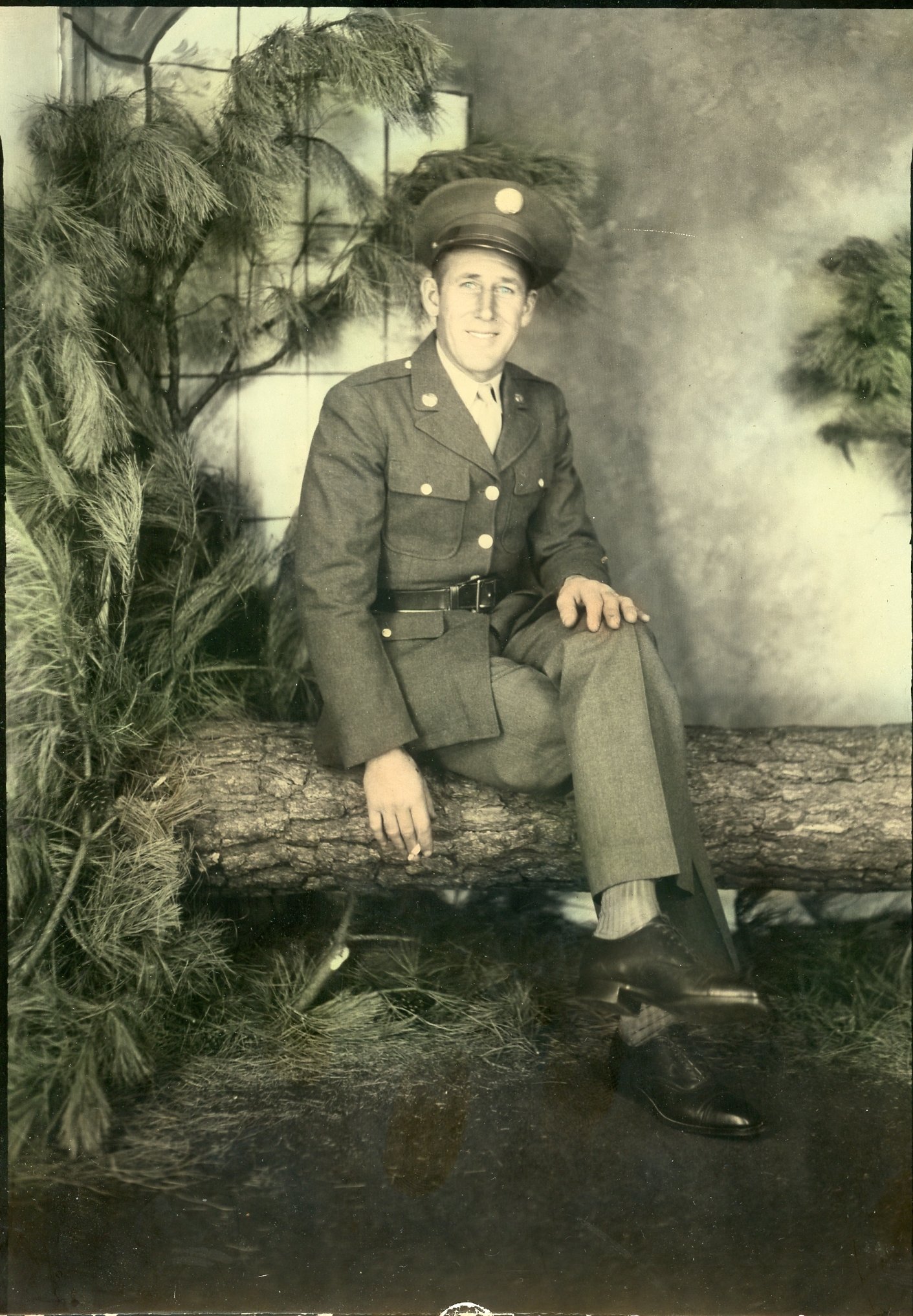 Vintage photo of a man in a military uniform sitting on a log, surrounded by foliage, with a neutral background.