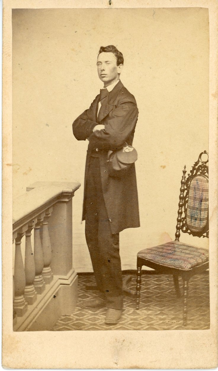 Vintage photo of a man in formal attire beside a carved chair with decorative upholstery.