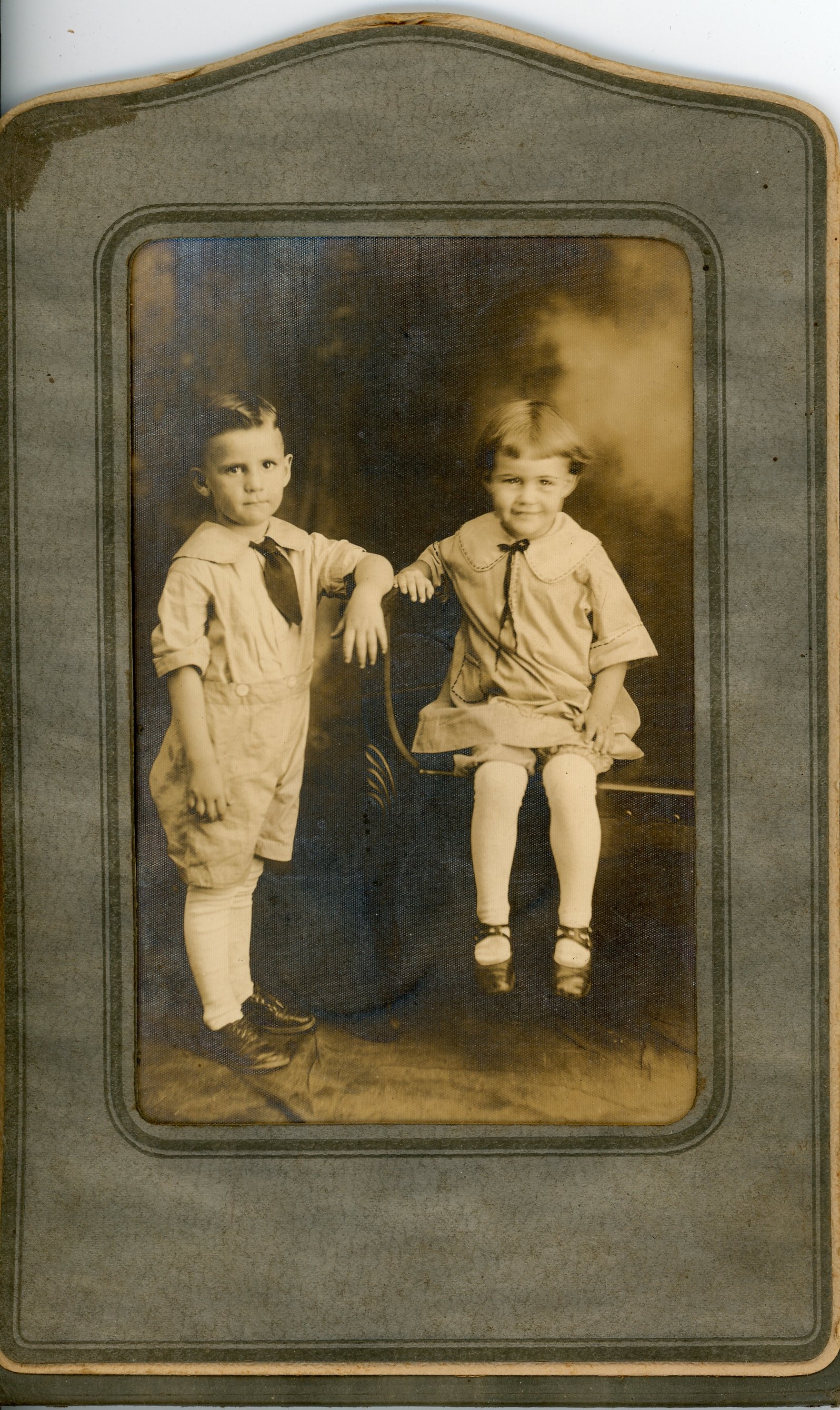 Vintage photo of two children; a boy in shorts and tie, and a girl in a dress and ribbon, posed and framed.