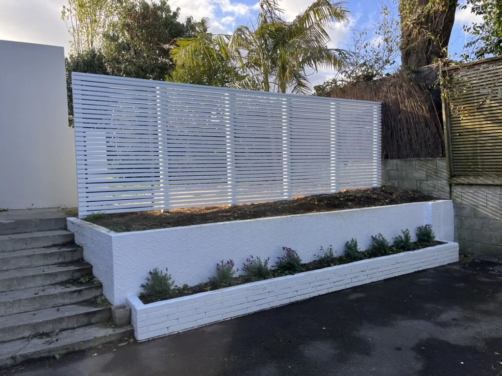 A backyard patio with a wooden bench, garden bed with plants, gravel area, and a curved concrete walkway, enclosed by a wooden privacy fence, under a partly cloudy sky.