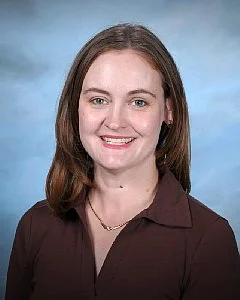 Portrait of a smiling woman with glasses and long gray hair against a light blue background.