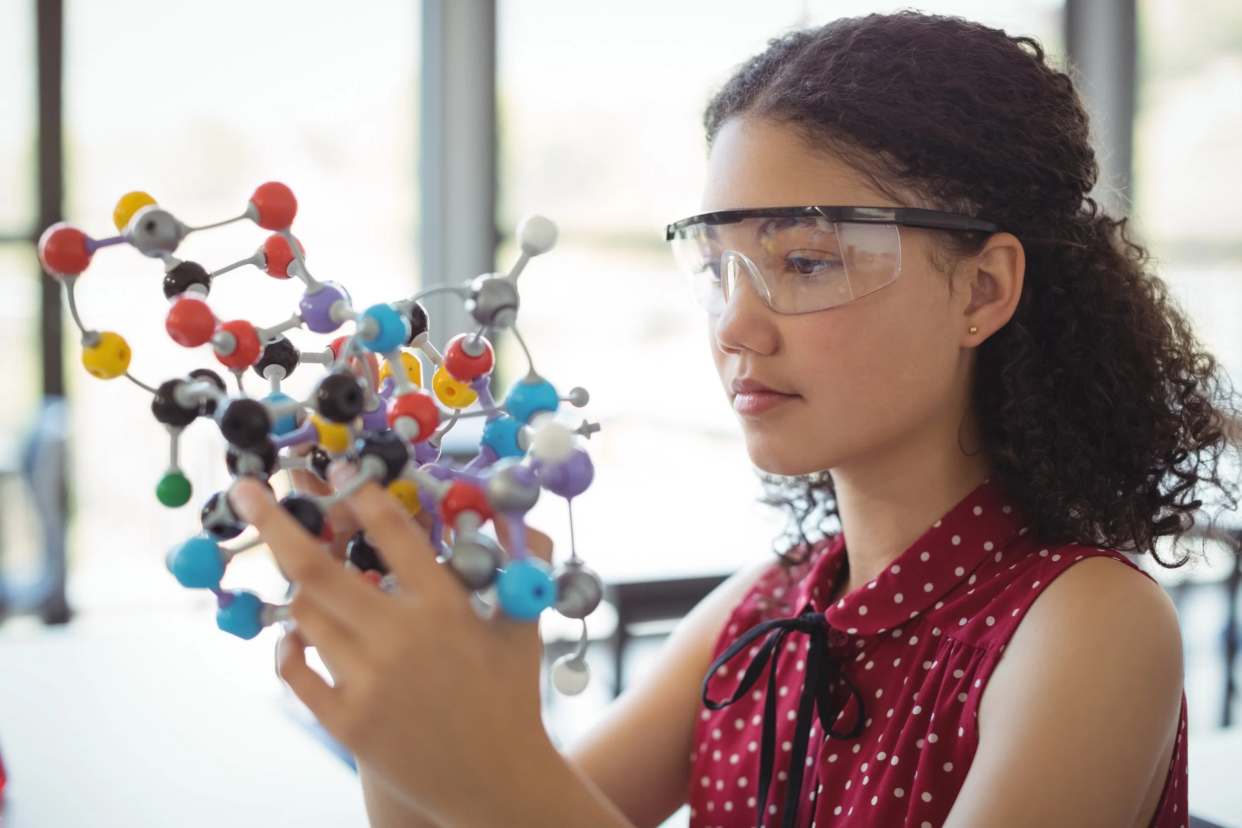 Female student wearing safety goggles examining a colorful molecular model in a classroom.