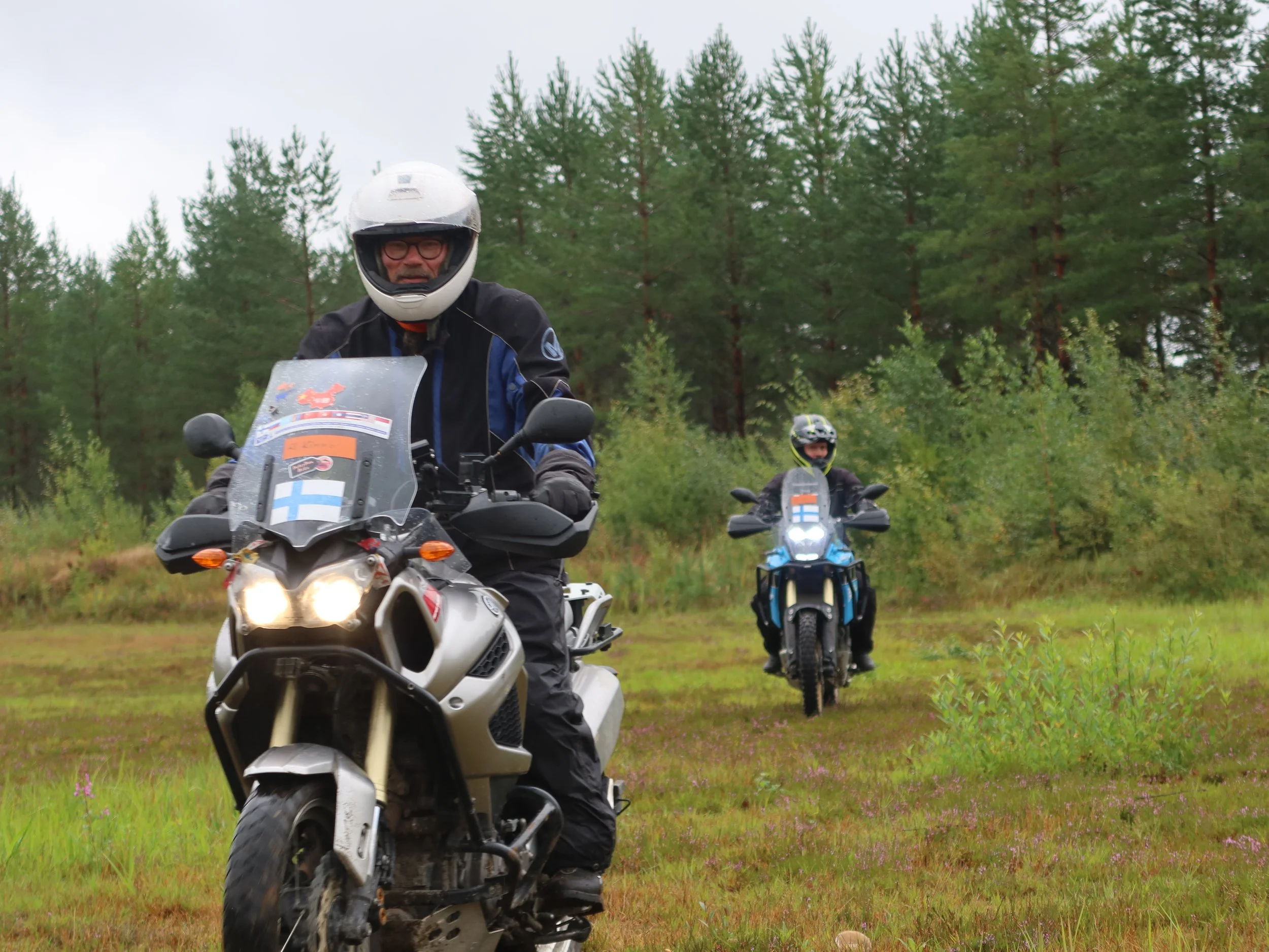 Kaksi mototrilaajia ajamassa metsän reunassa, molemmilla kypärä ja suojavaatteet, etummainen Suomen lipputarra ja keltainen numero. Taustalla puita ja vihreitä pensaita.