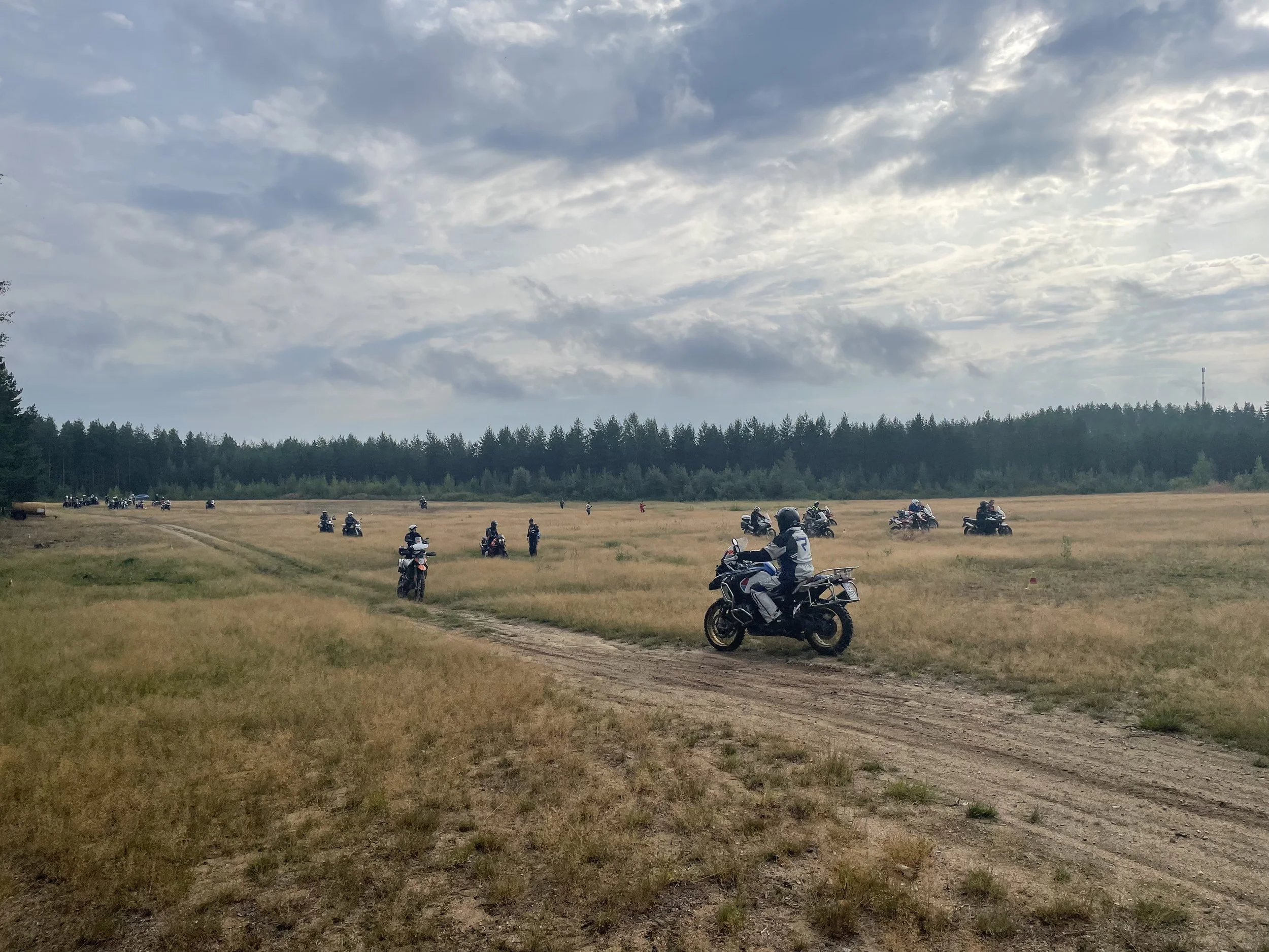 Moottoripyöriä ja motoristeja maaseudulla, pilvinen taivas ja metsäinen tausta.