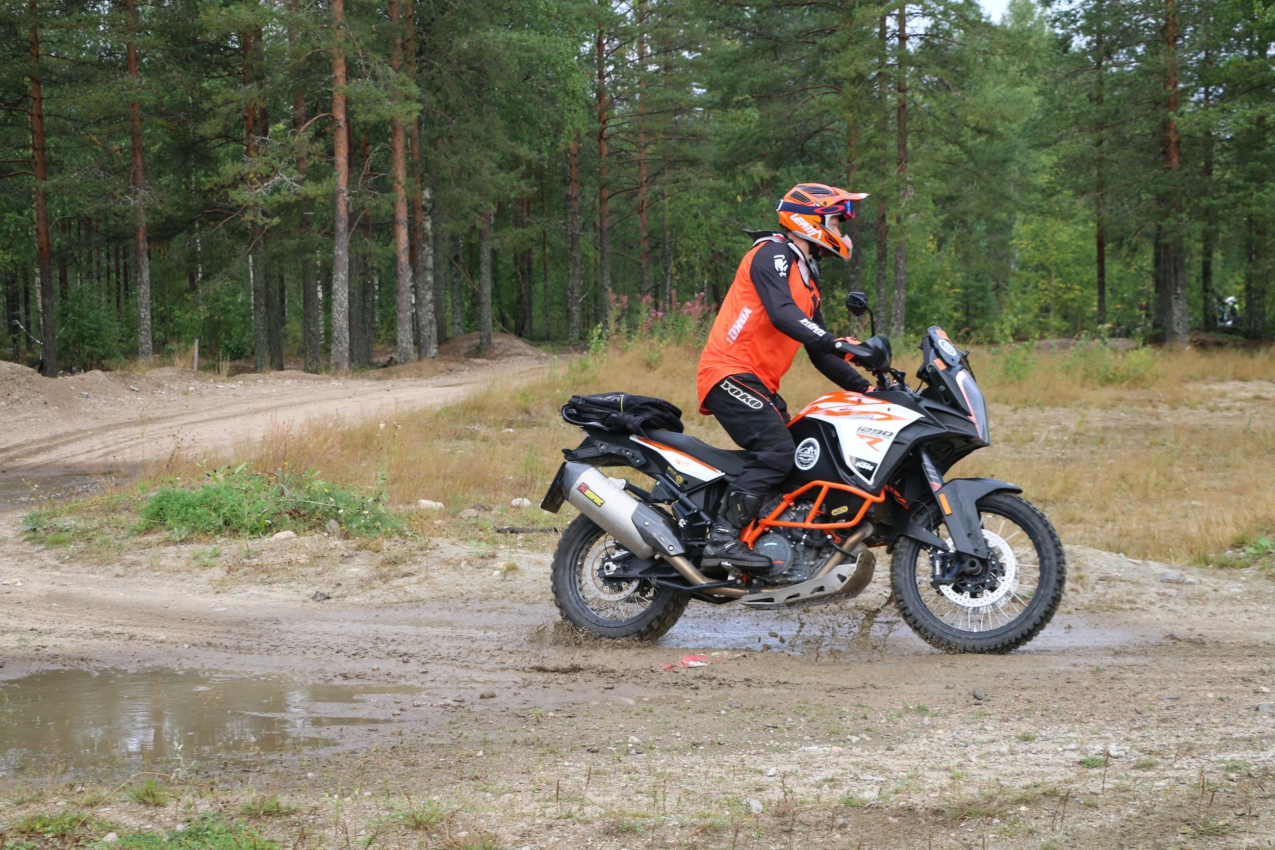 Moottoripyöräilijä ajamassa metsän maastossa, tietä pitkin, asusteet ja pyörä ovat oranssin ja mustan väriset.