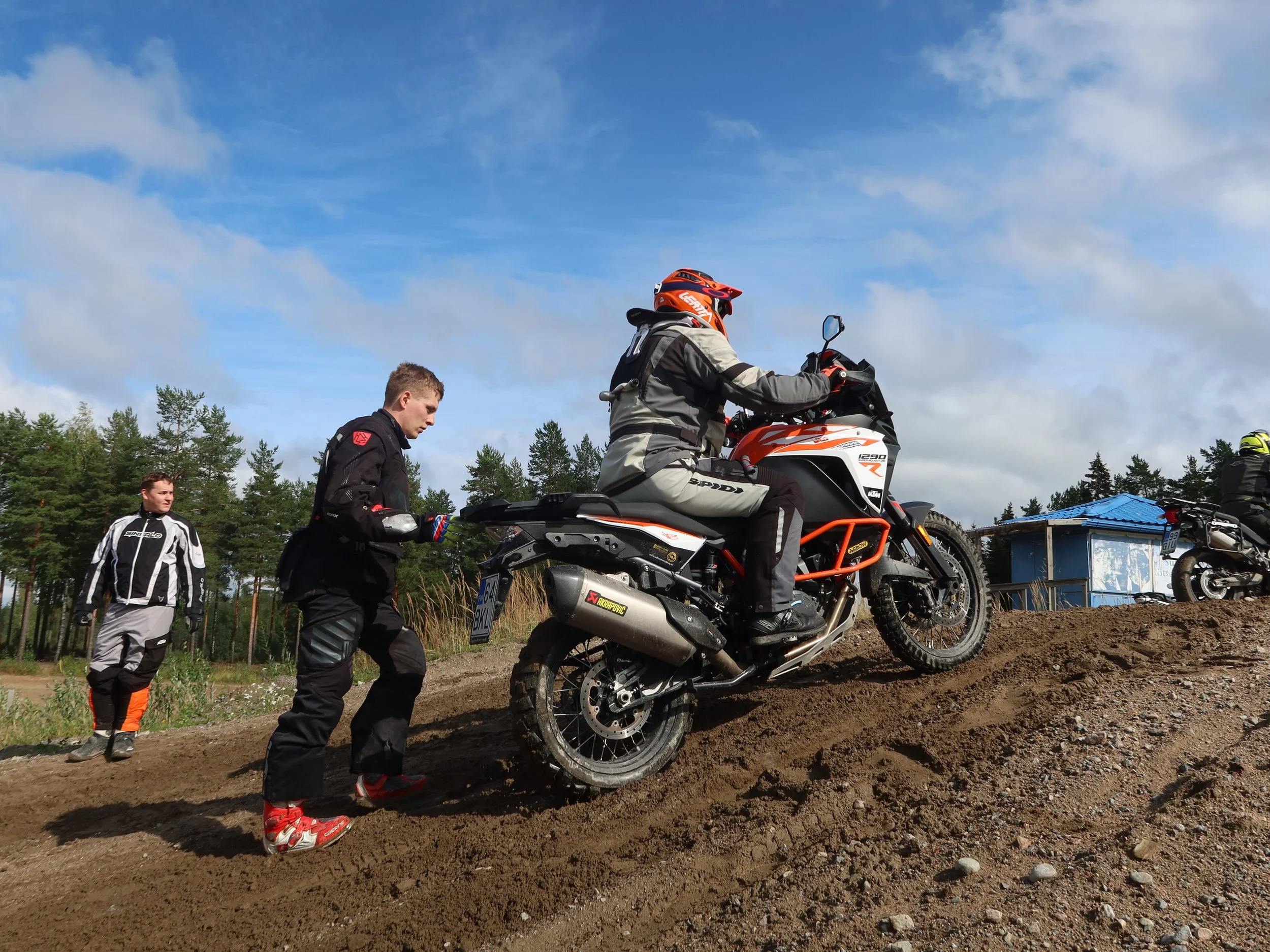 Mies polkee maastoajoneuvon ylös mäkeä. Hän on moottoripyöräilijä ja puetaan suojavarusteisiin. Taustalla kolme muuta moottoripyöräilijää ja metsää.