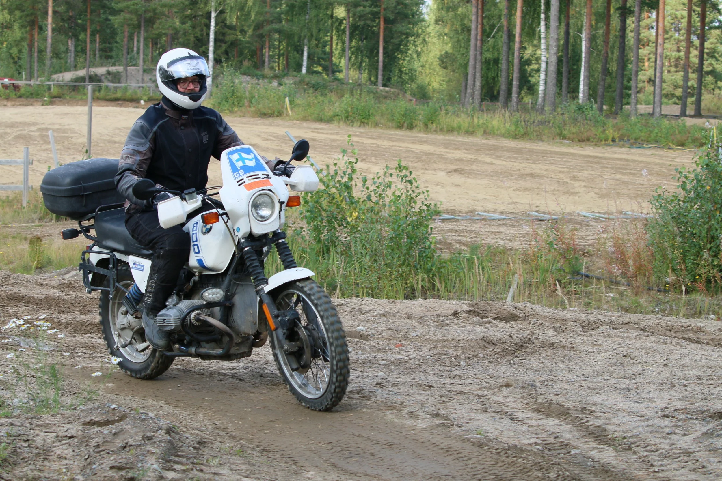 Mies moottoripyörällä ratsastamassa polulla, taustalla metsä ja avoin maa-alue.