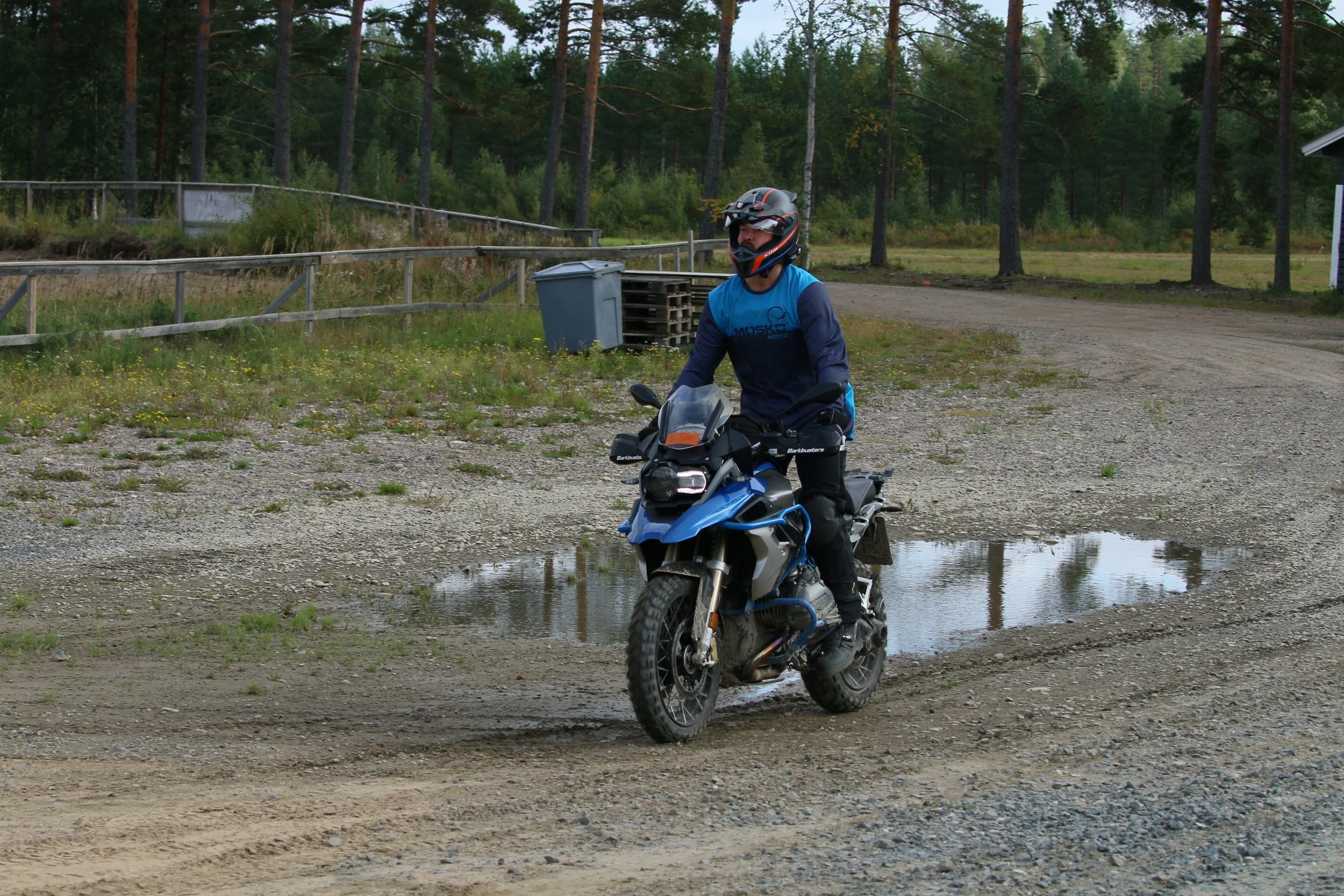 Mies ratsastaa moottoripyörällä pojat maastossa, jalaseinä ja puita taustalla.