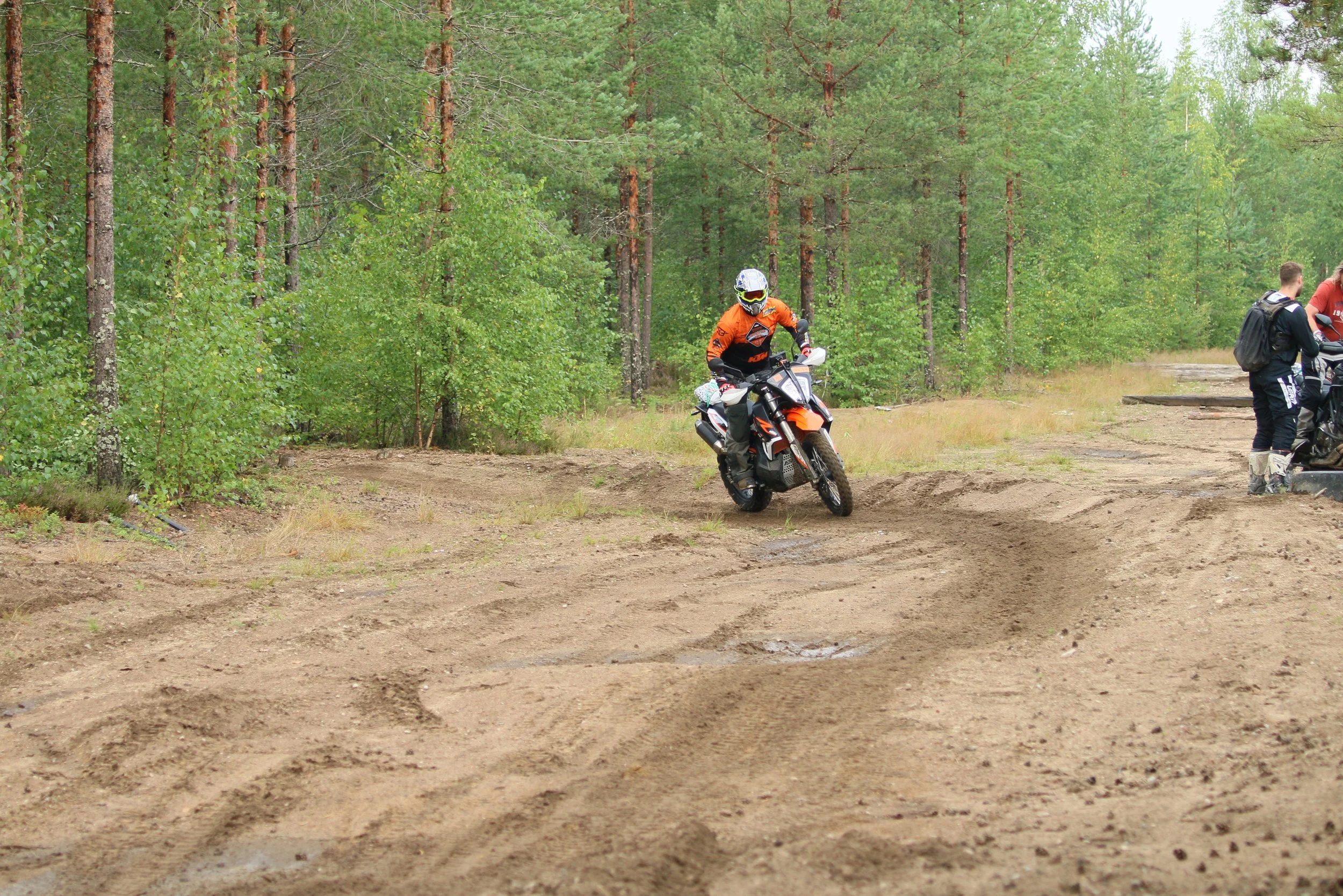 Kuljettaja ajaa motocross-moottoripyörää metsäisellä polulla, ympärillä puita ja kaksi muuta ihmistä seisomassa reitin reunalla.